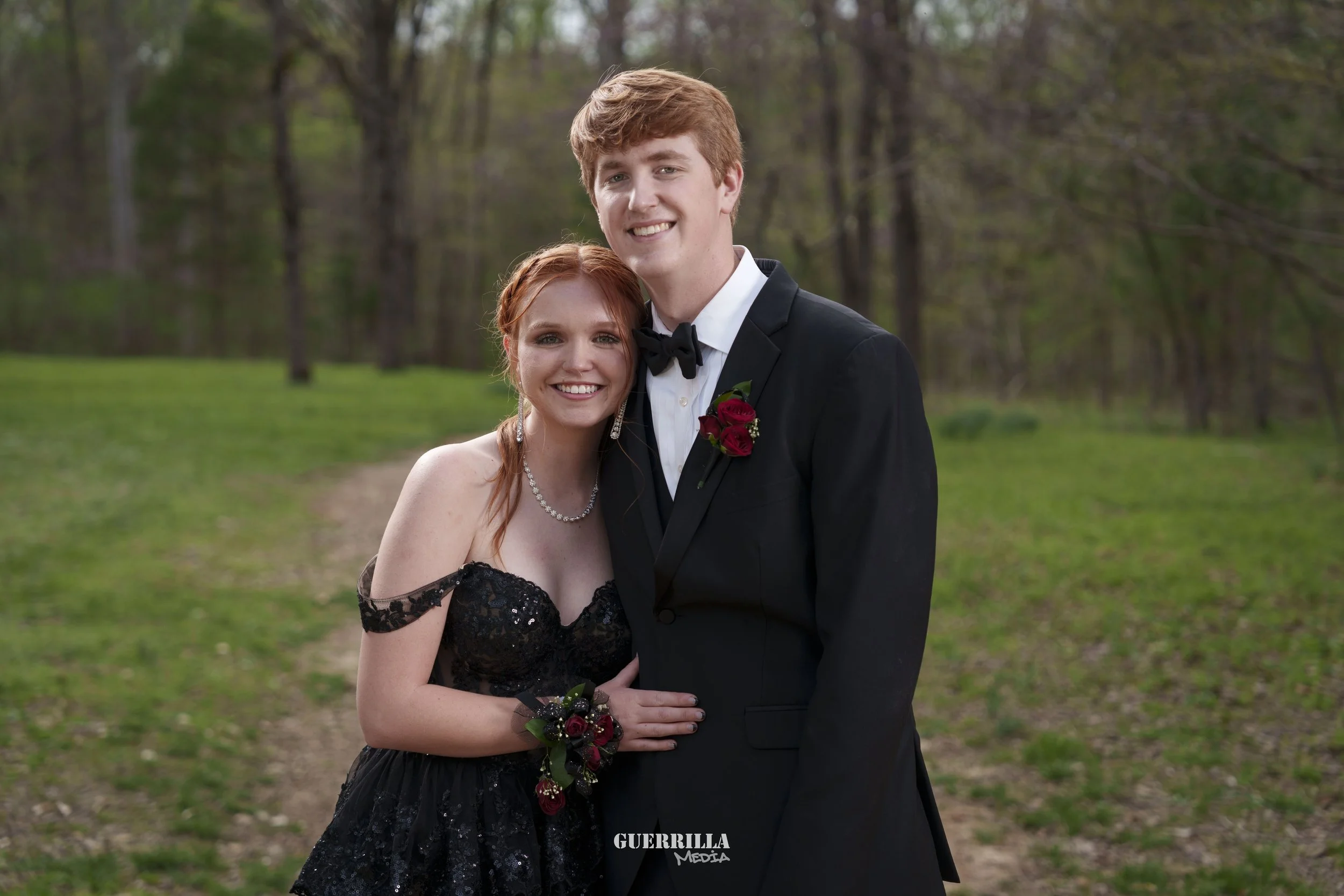 Young couple in formal attire standing outdoors, smiling, with trees and green grass in the background.