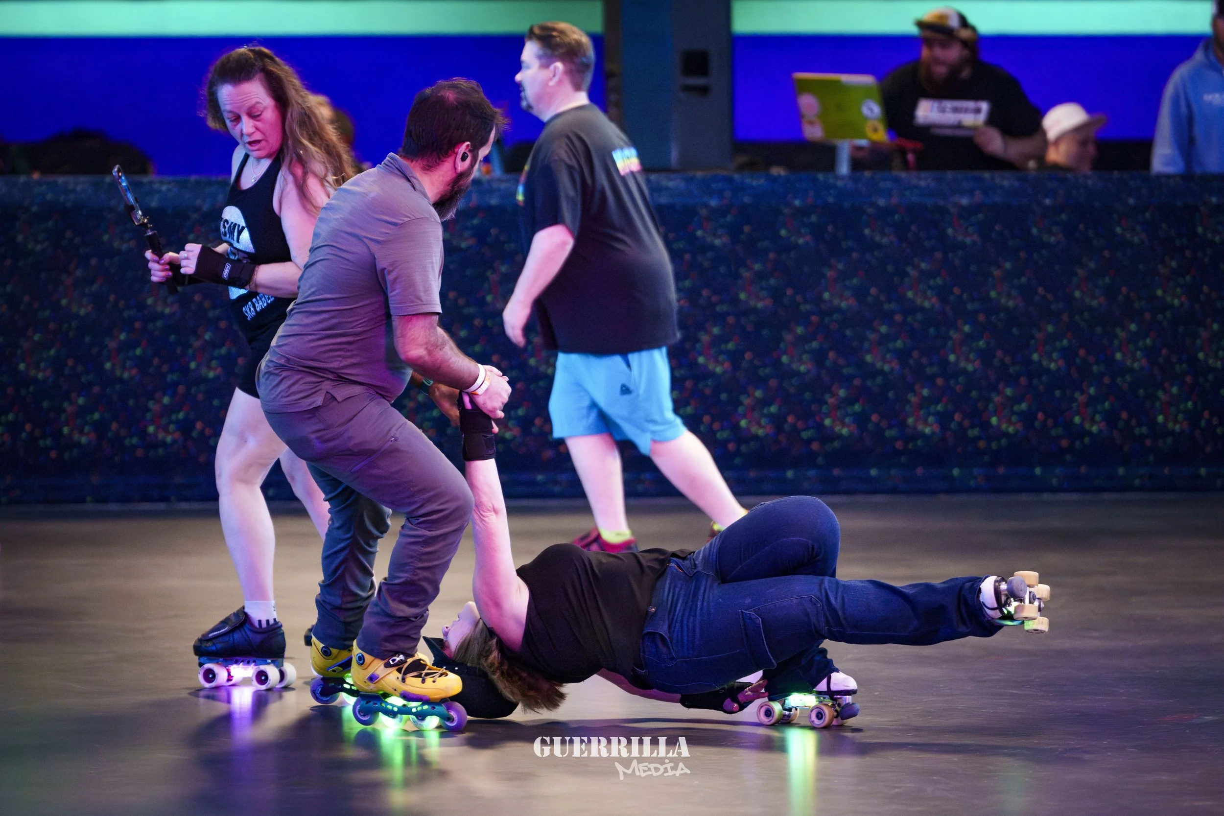 Two people helping a woman who fell while roller skating on a rink. There are additional people in the background, and the scene is indoors with colorful lighting.