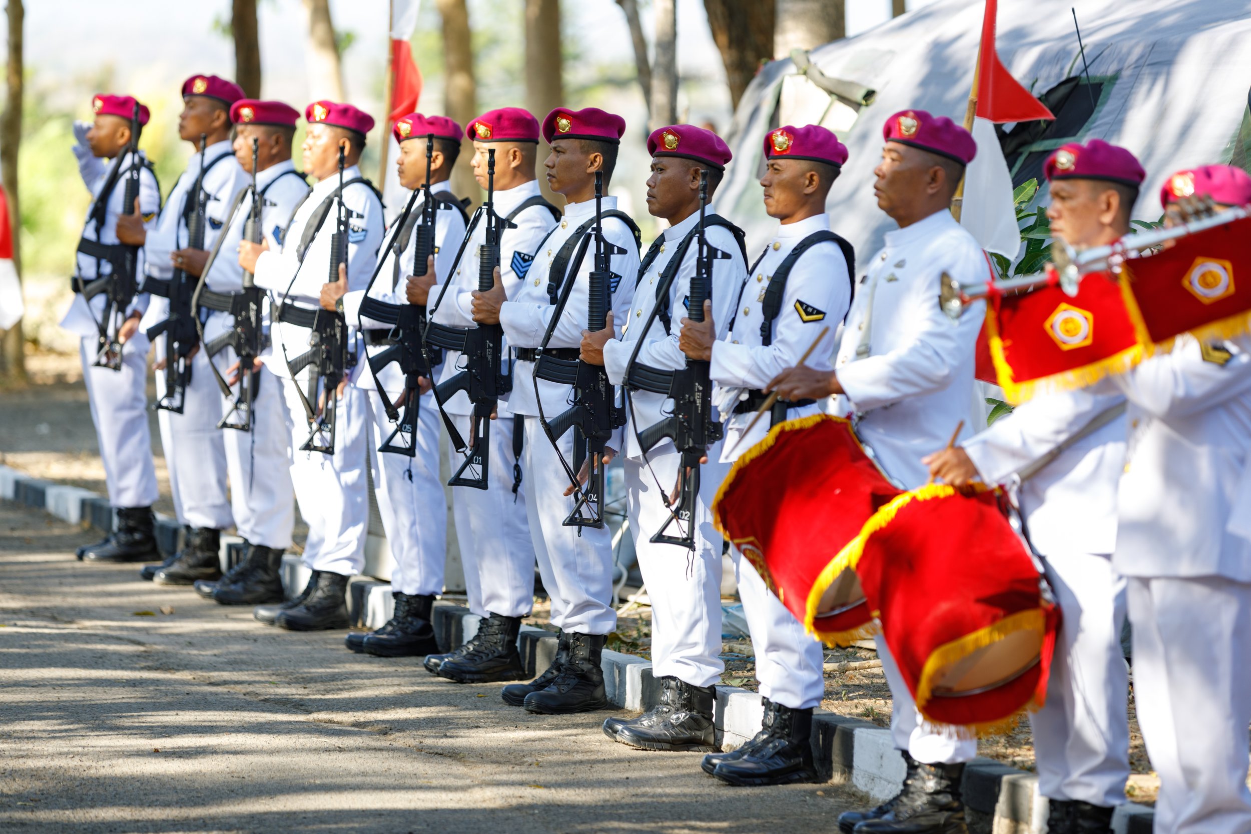A line of soldiers in white uniforms and maroon berets standing at attention holding rifles, with a drummer in white uniform and maroon and red drum playing nearby, during a parade or ceremony outdoors.