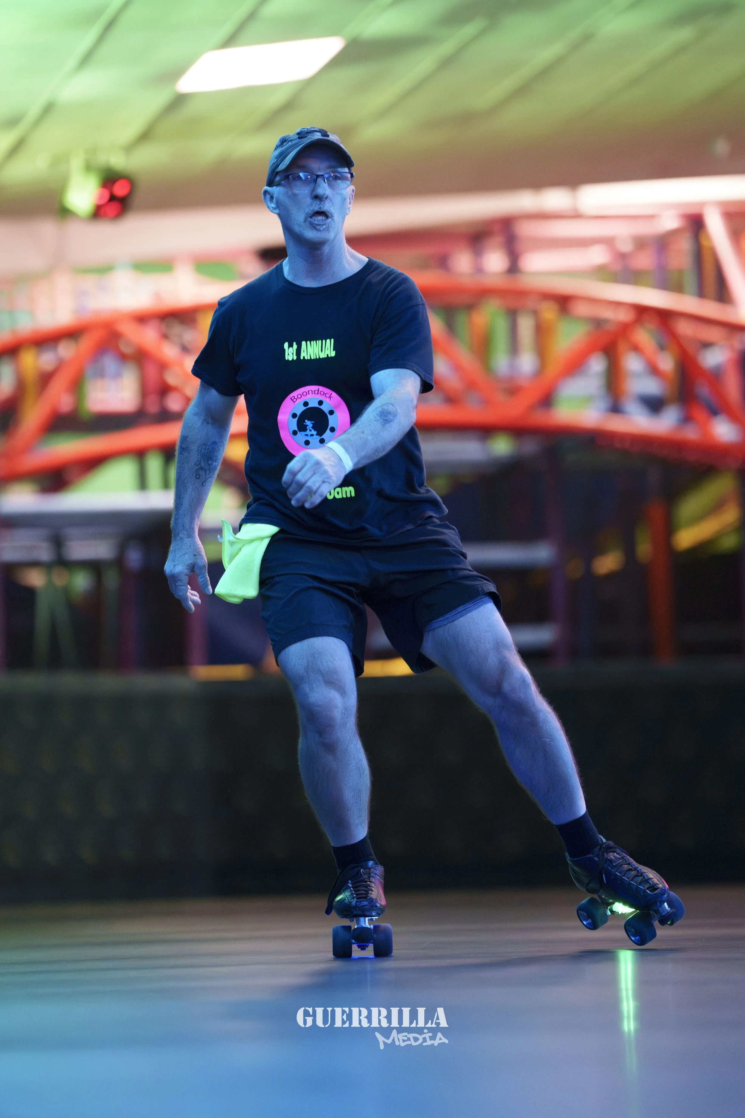 A man roller skates indoors wearing glasses and a black t-shirt with neon green and pink lettering, and a black cap.