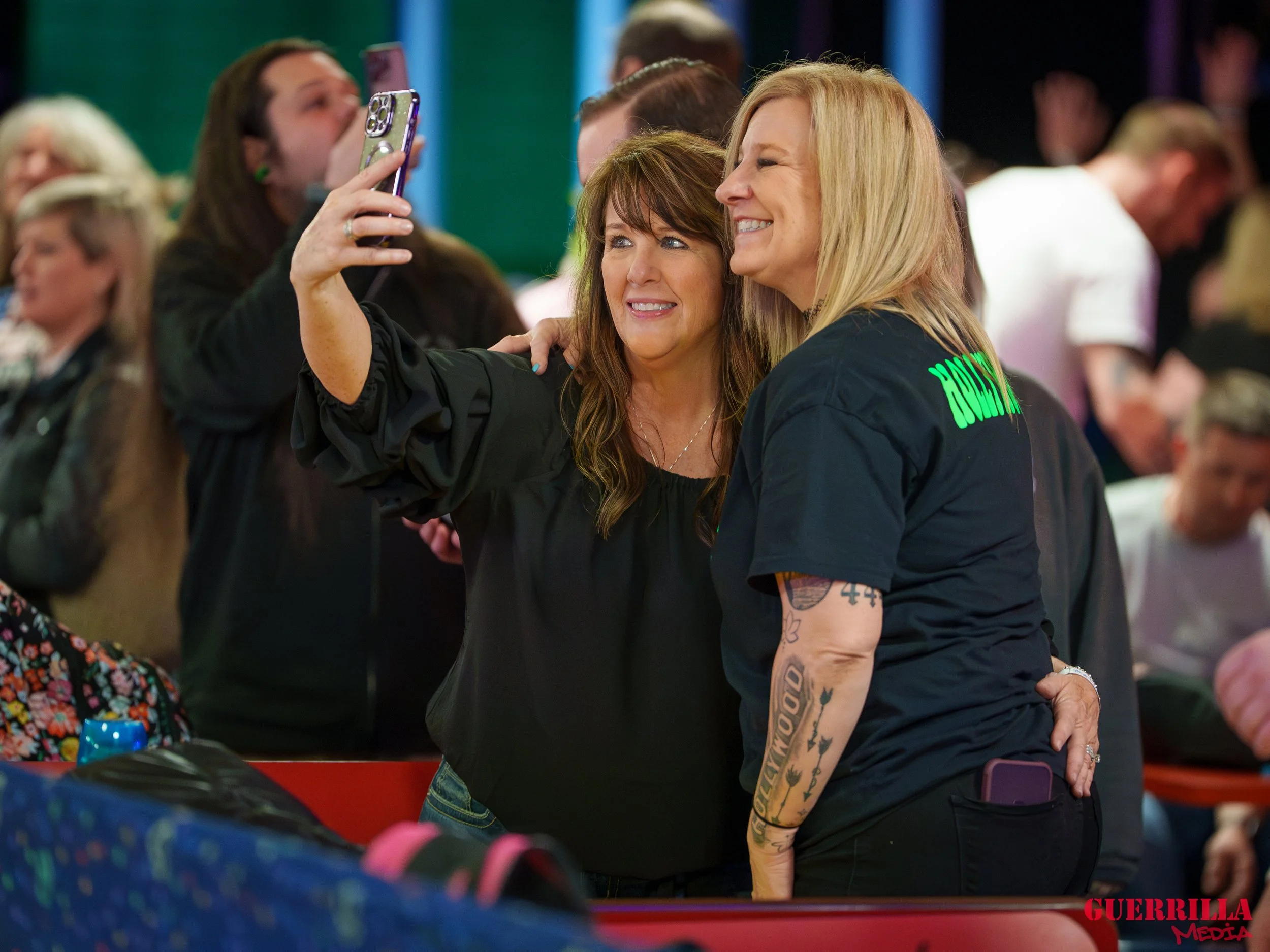 Two women taking a selfie at a crowded indoor event, smiling and close together, with other people in the background.