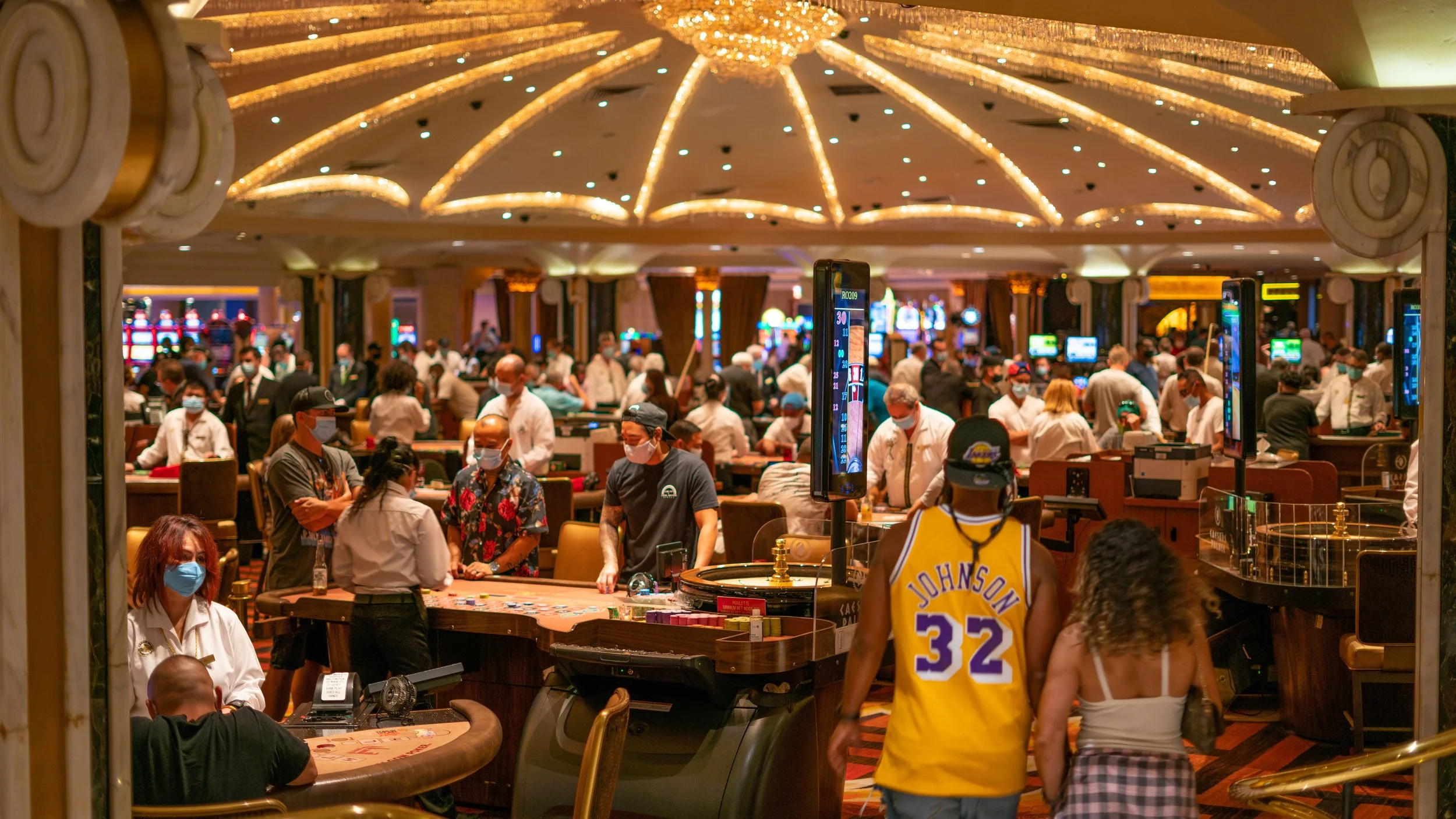 Inside a casino with many people playing and gambling, some wearing masks, and a young woman in a Lakers jersey walking with a woman in a white top.