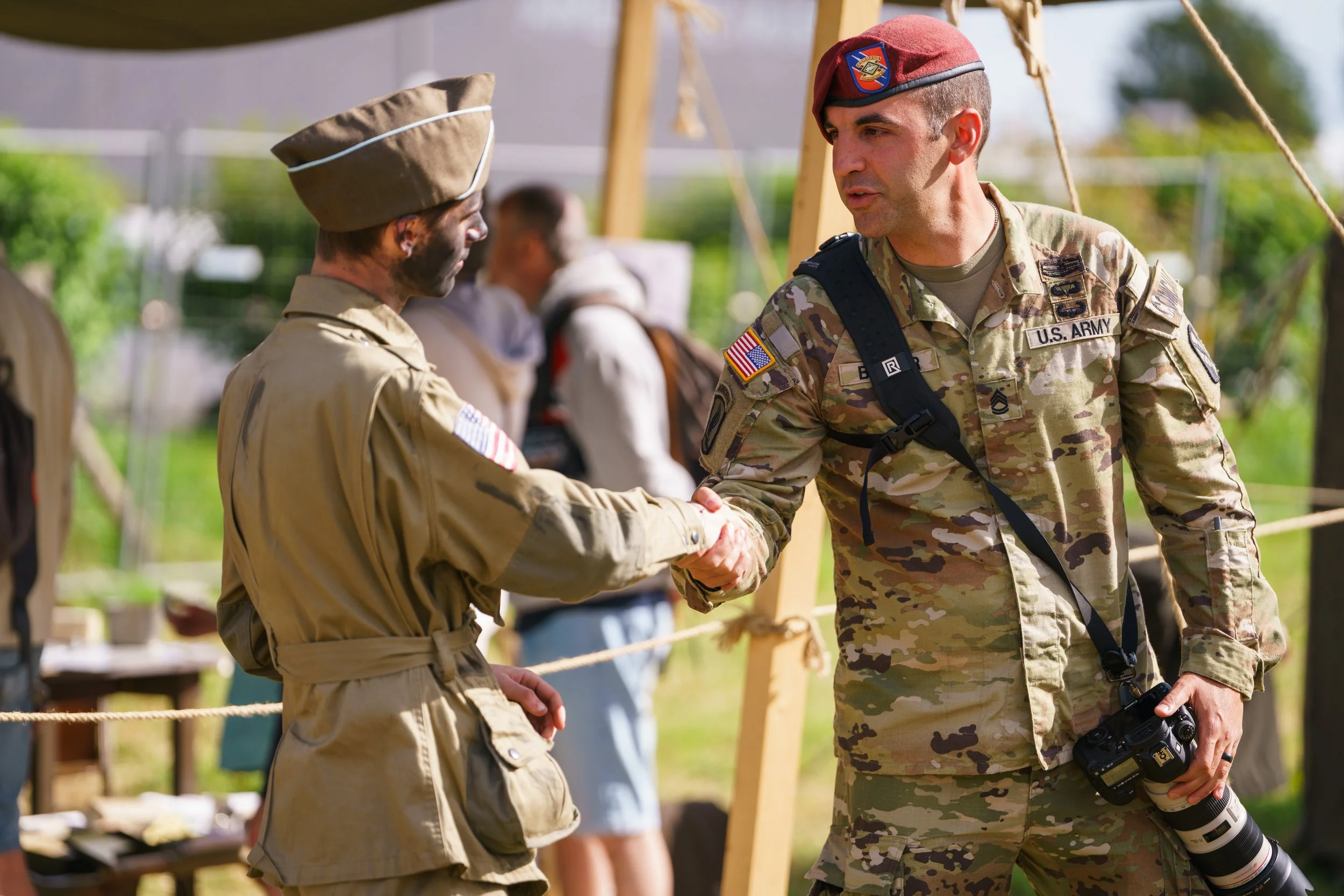 Two soldiers, one in a tan uniform and the other in camouflage, shake hands outdoors during a military event. The soldier in camouflage holds a camera with a large lens and has a U.S. Army patch. The background shows other people and event setup.