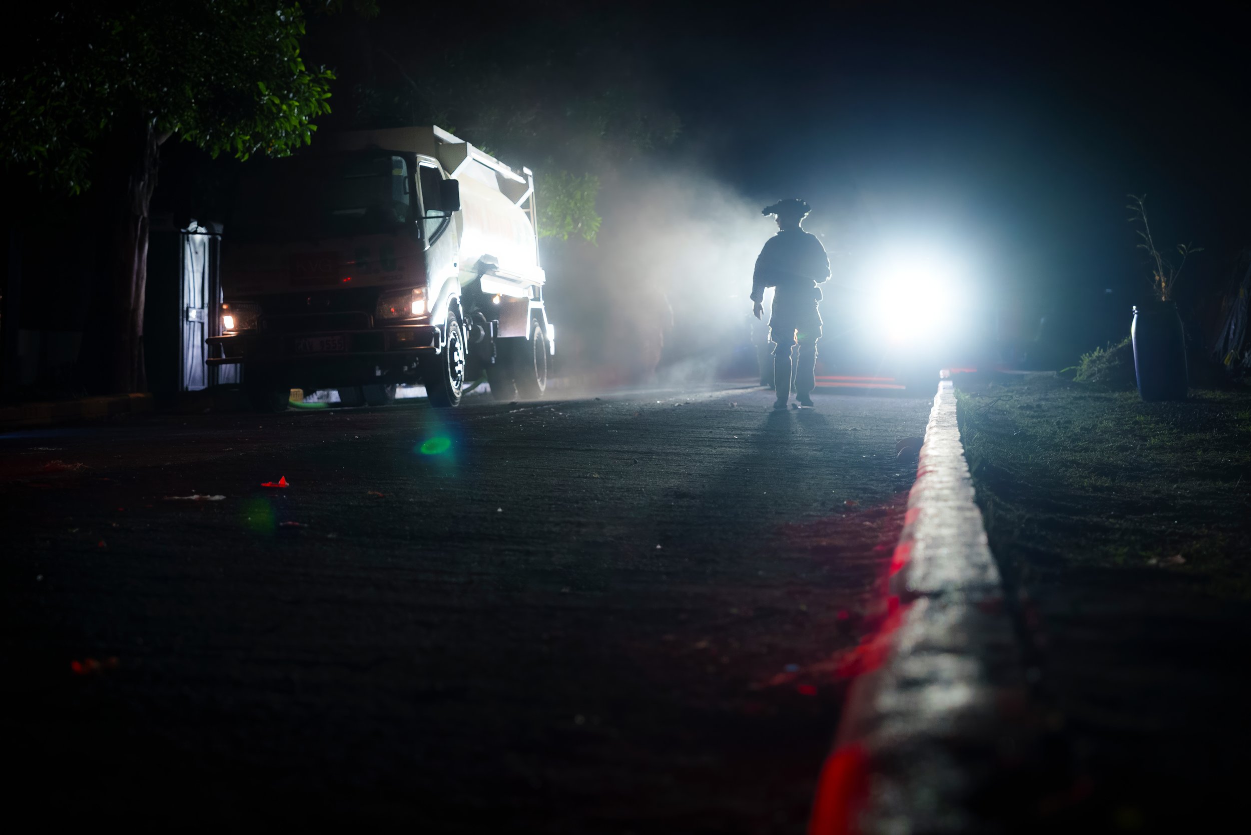 A Soldier walking on a dark street at night with a bright light behind him, a truck nearby, and smoke in the air.