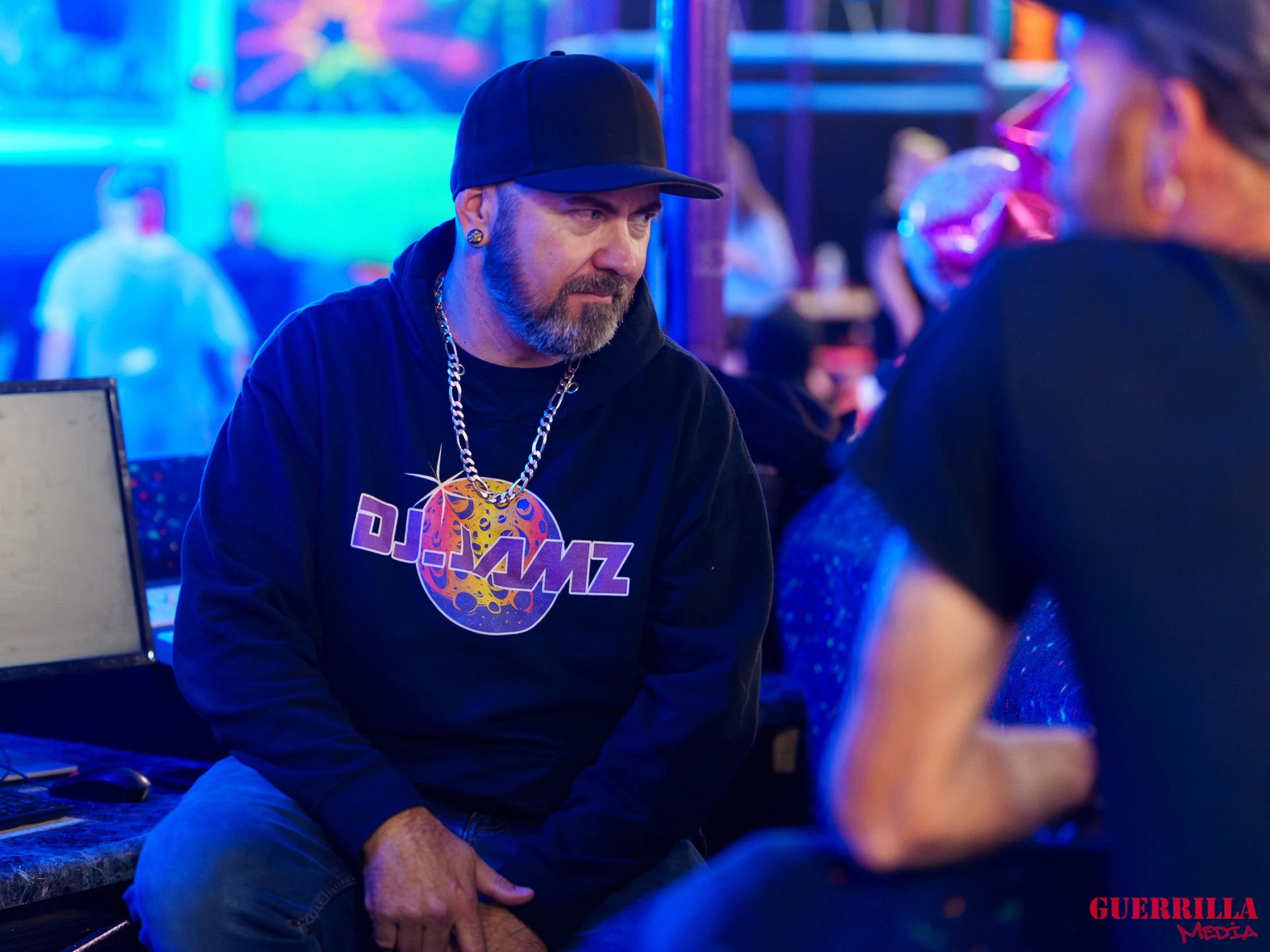A man with a beard and earrings, wearing a black cap and a black hoodie with a "DJ JAZZ" logo and a large chain necklace, is sitting in a nightclub or bar, engaged in conversation with someone whose back is to the camera. The background is colorful w