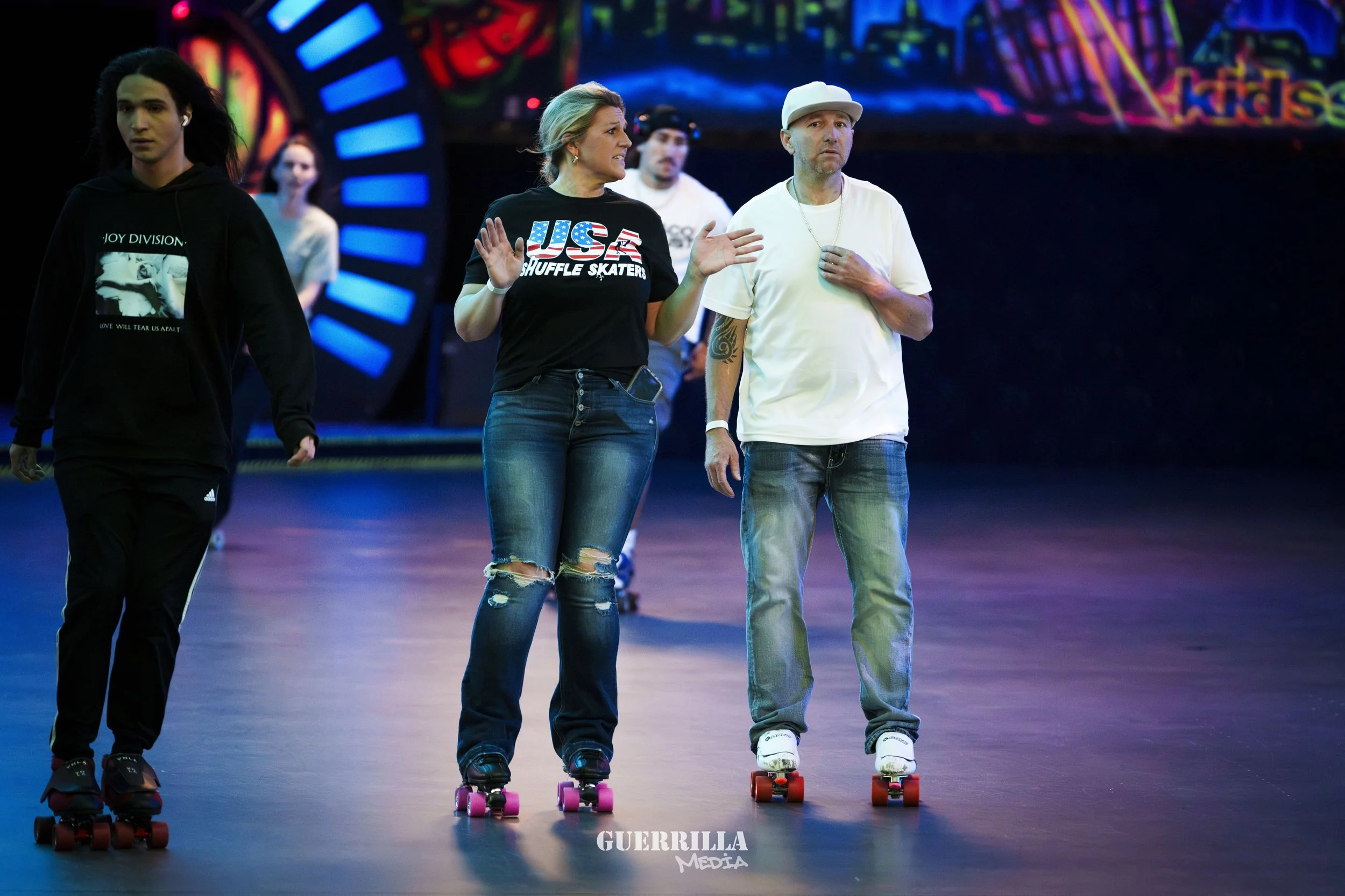 People roller skating in an indoor rink with colorful background lights and a large illuminated clock.
