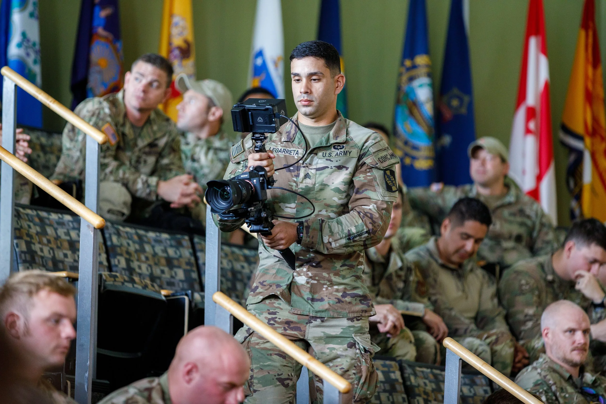 A soldier in camouflage uniform holding a camera, surrounded by other soldiers sitting and listening, with several flags in the background.