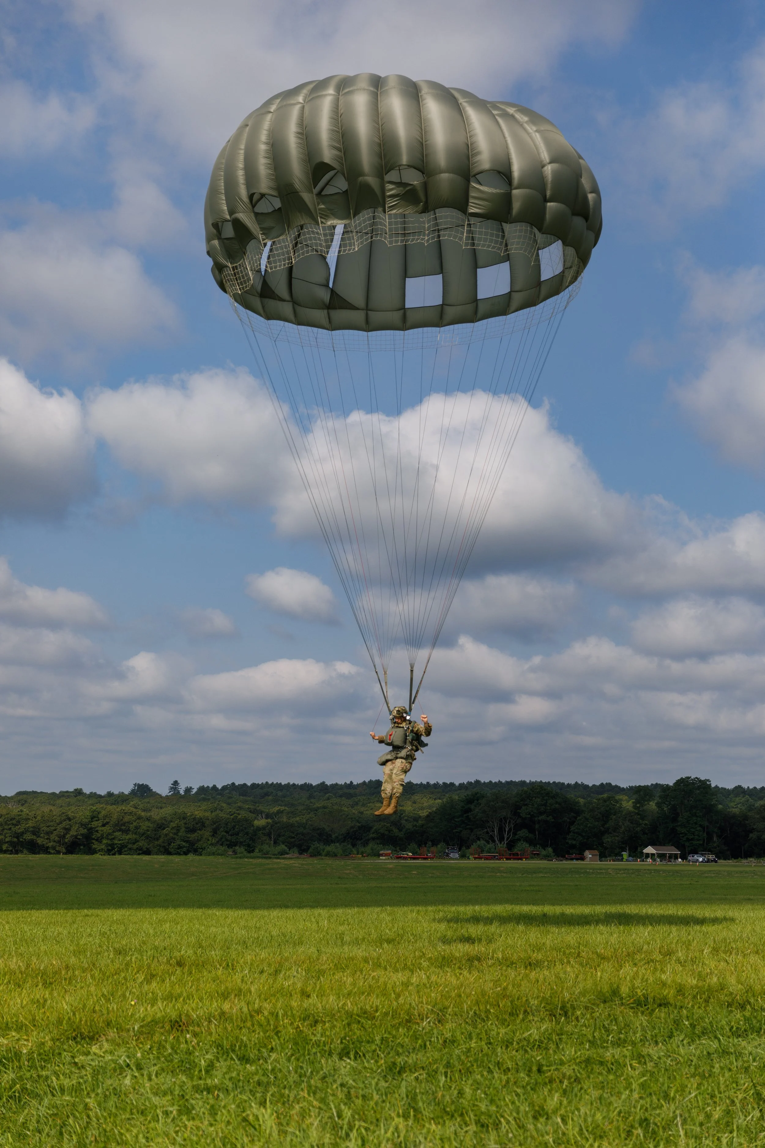 A soldier is parachuting down with a dark green parachute over a green field and a cloudy sky.