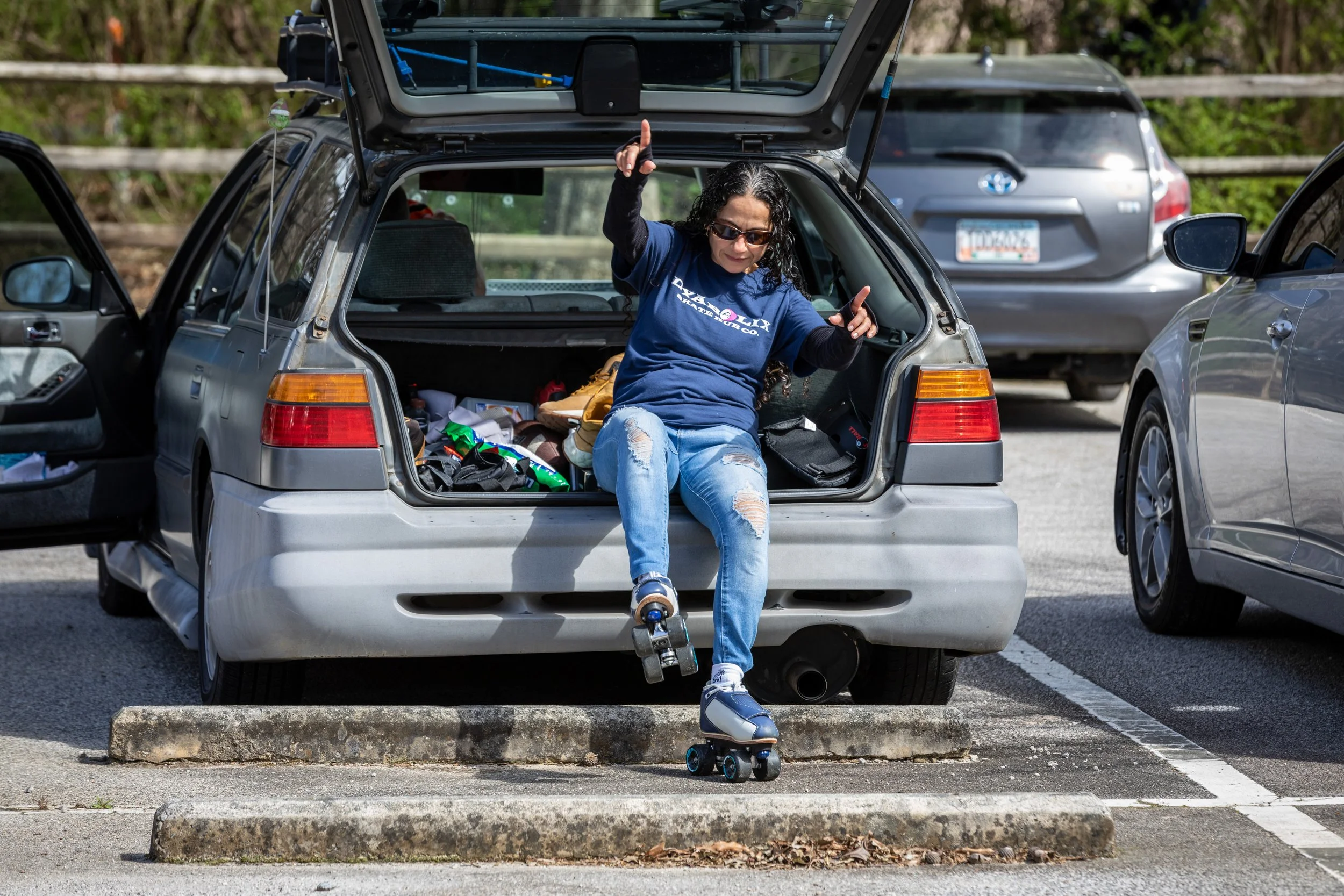 Woman sitting in the open trunk of a silver car, wearing roller skates and ripped jeans, gesturing with both hands, and smiling. The trunk contains various items, and the car is parked in a parking lot.