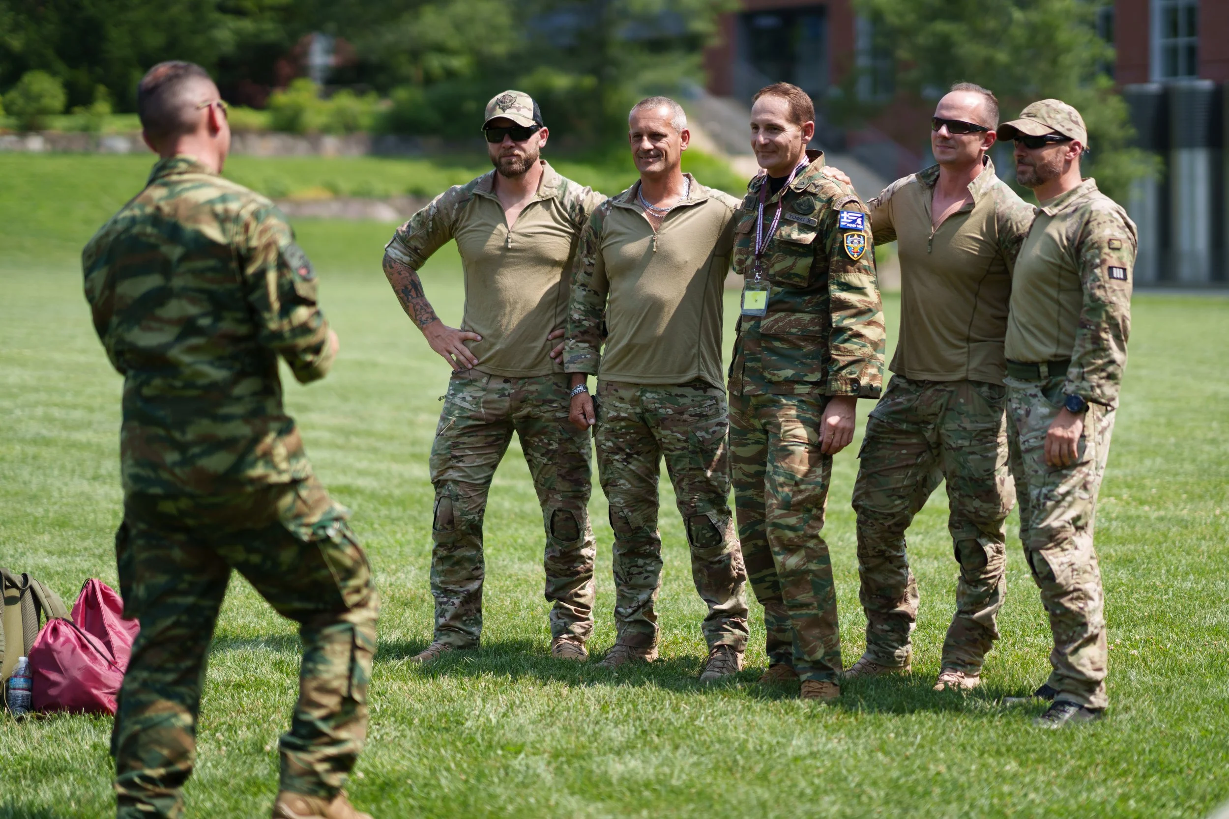 Group of soldiers in camouflage uniforms standing on grass while another soldier addresses them outdoors on a sunny day.