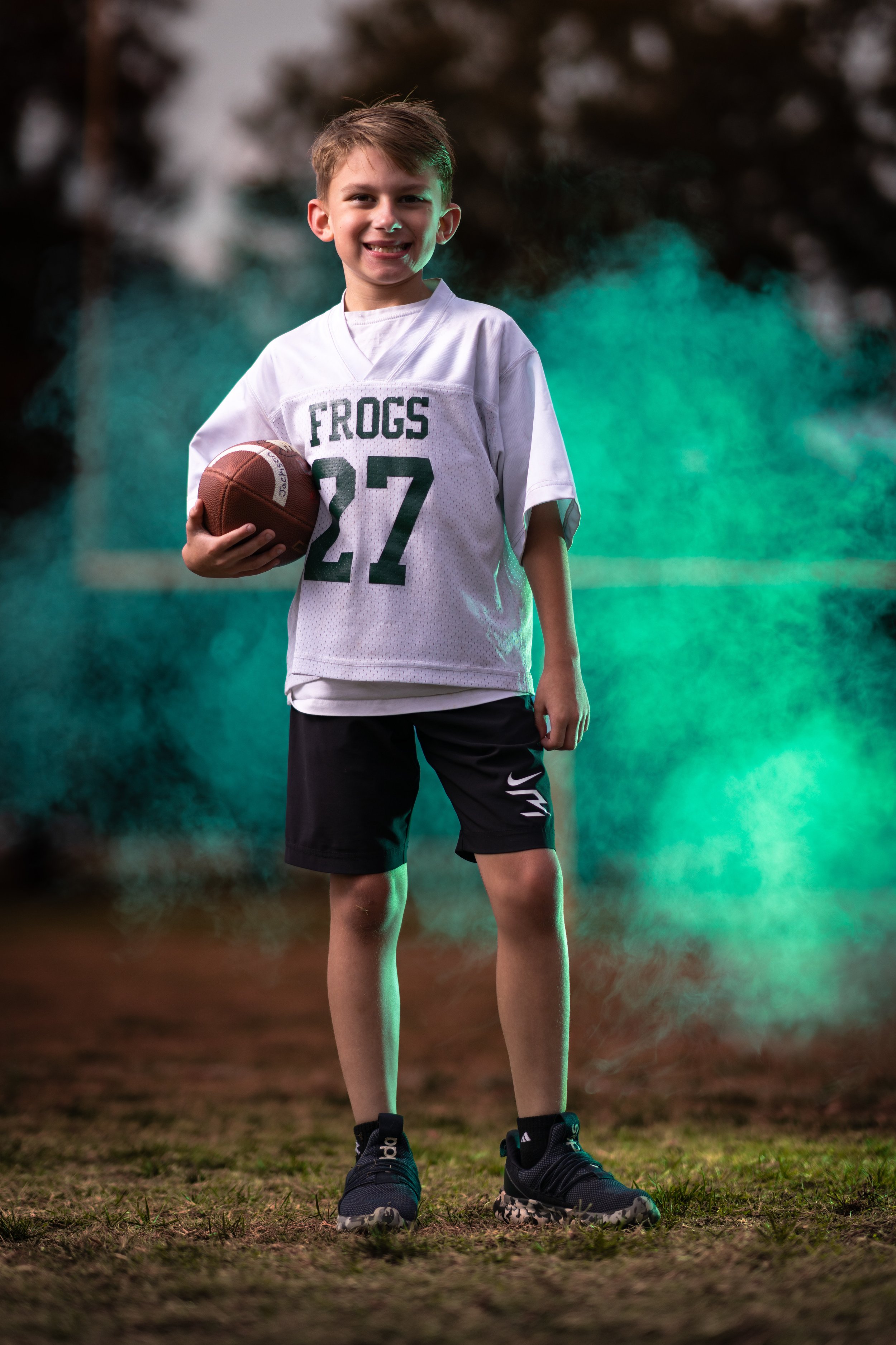 Young boy in a football uniform holding a football on a field with green and blue smoke in the background.