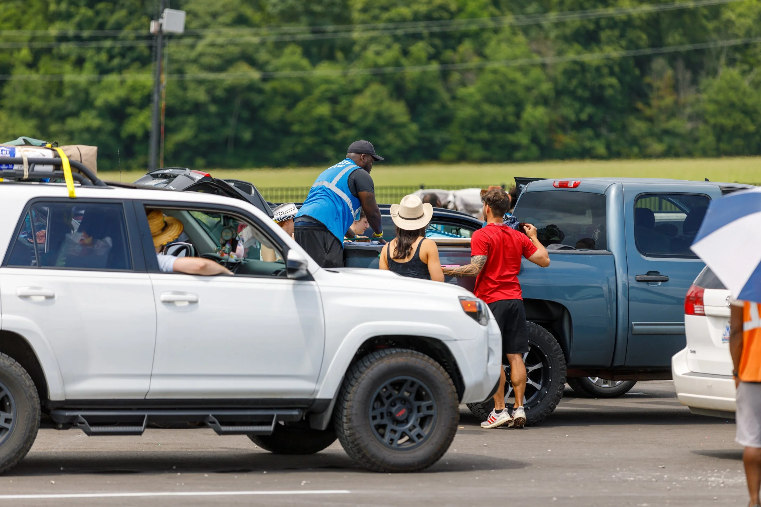 People gathered around vehicles at an outdoor event on a sunny day, with trees in the background.