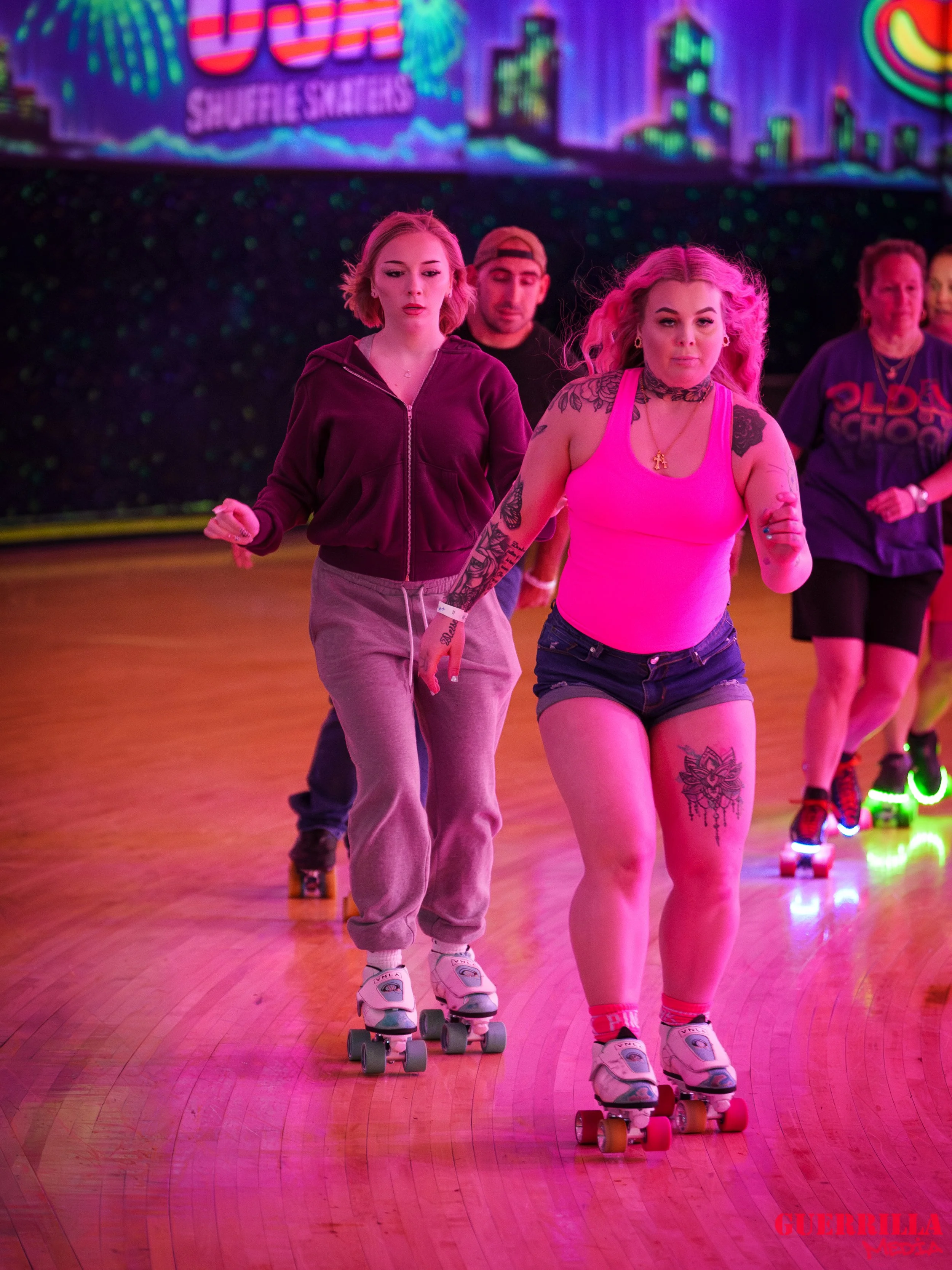 Group of people roller skating on a brightly lit indoor skating rink. The background features a neon sign with colorful graphics.