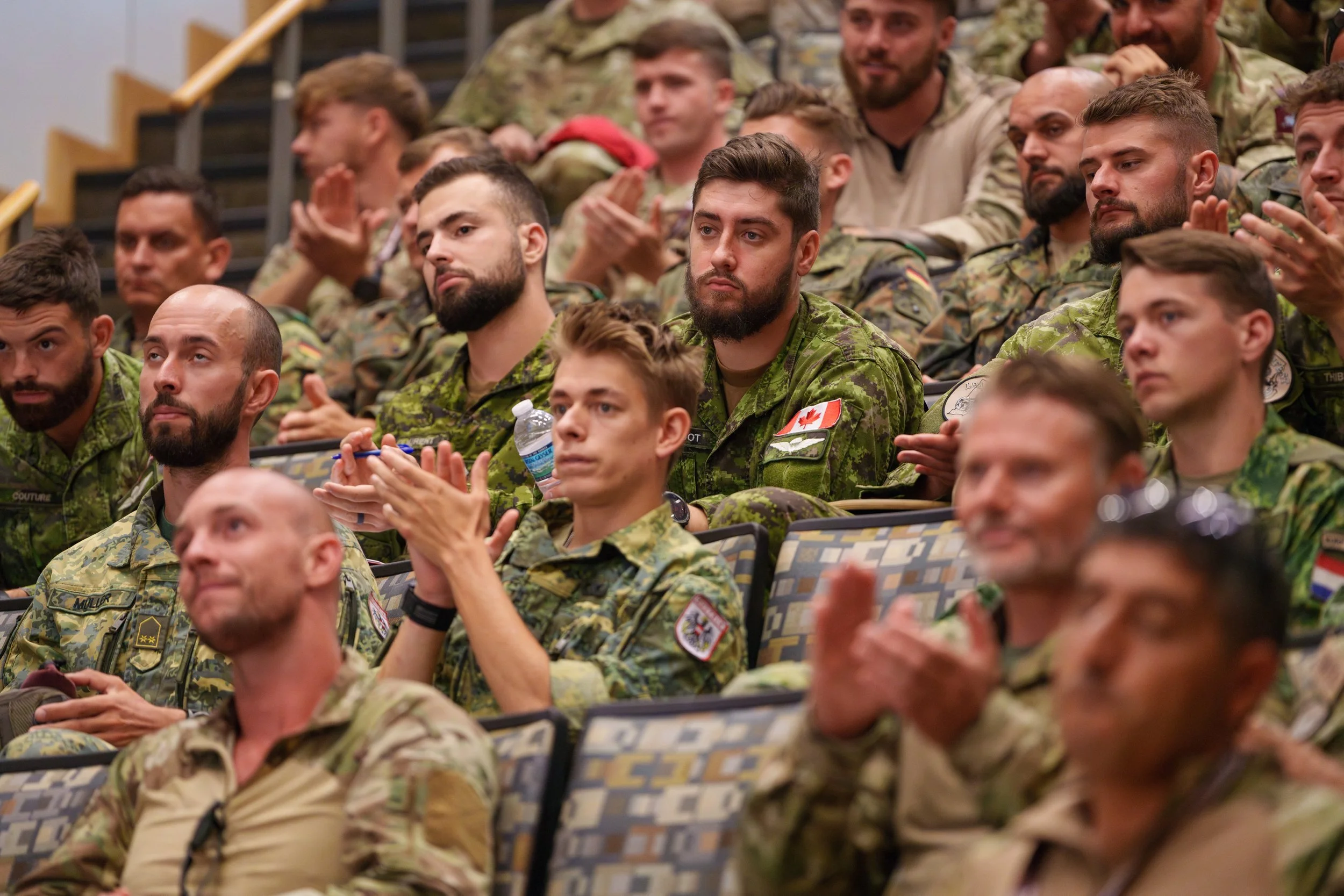 A group of soldiers, many with Canadian military patches, sitting in an auditorium or hall, paying attention and clapping, some holding water bottles.
