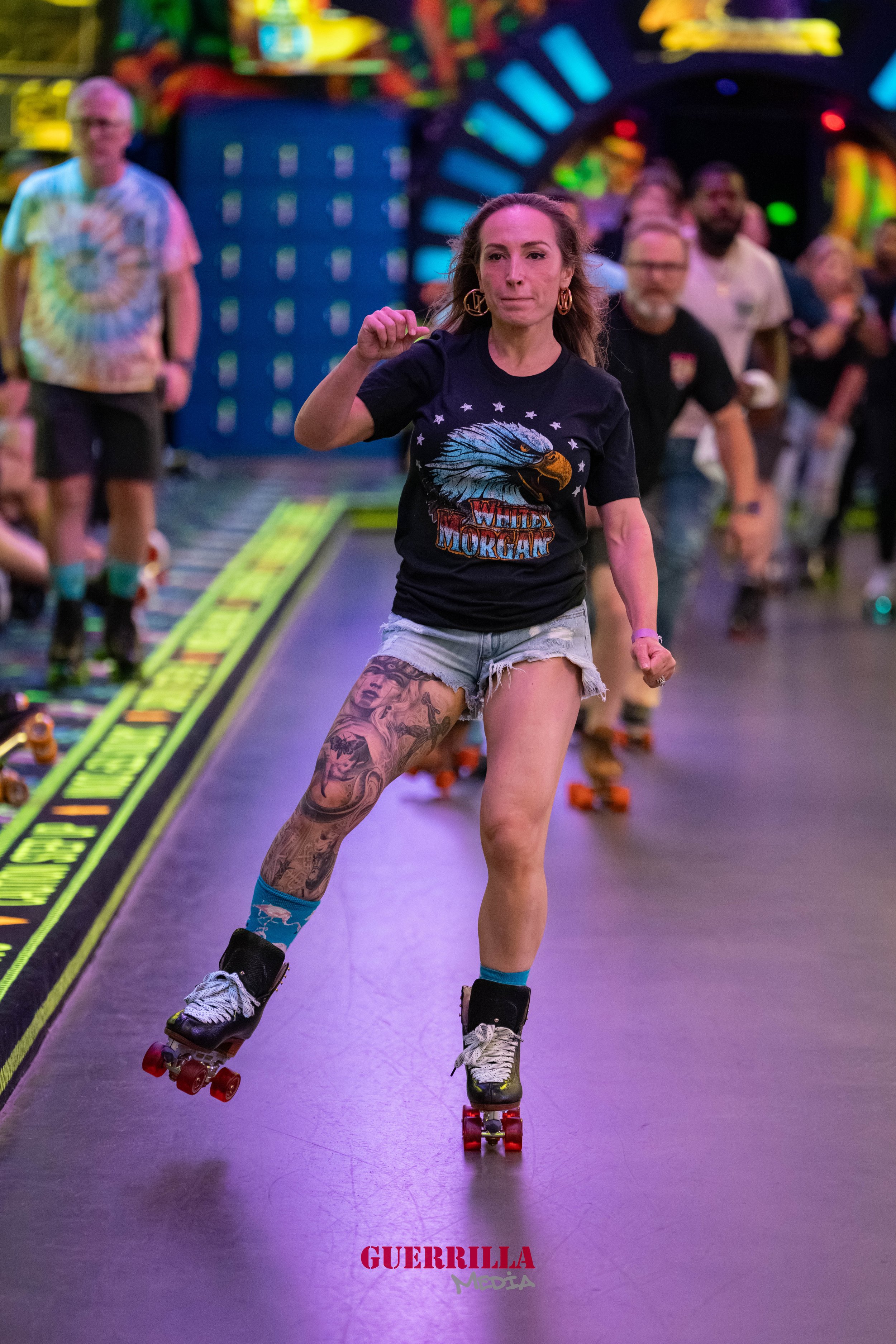 A woman roller skating indoors at a skate rink, wearing a black T-shirt with an eagle graphic and 'Willy Morgan' text, ripped denim shorts, and tattoos on her leg. She is surrounded by people and colorful lights.