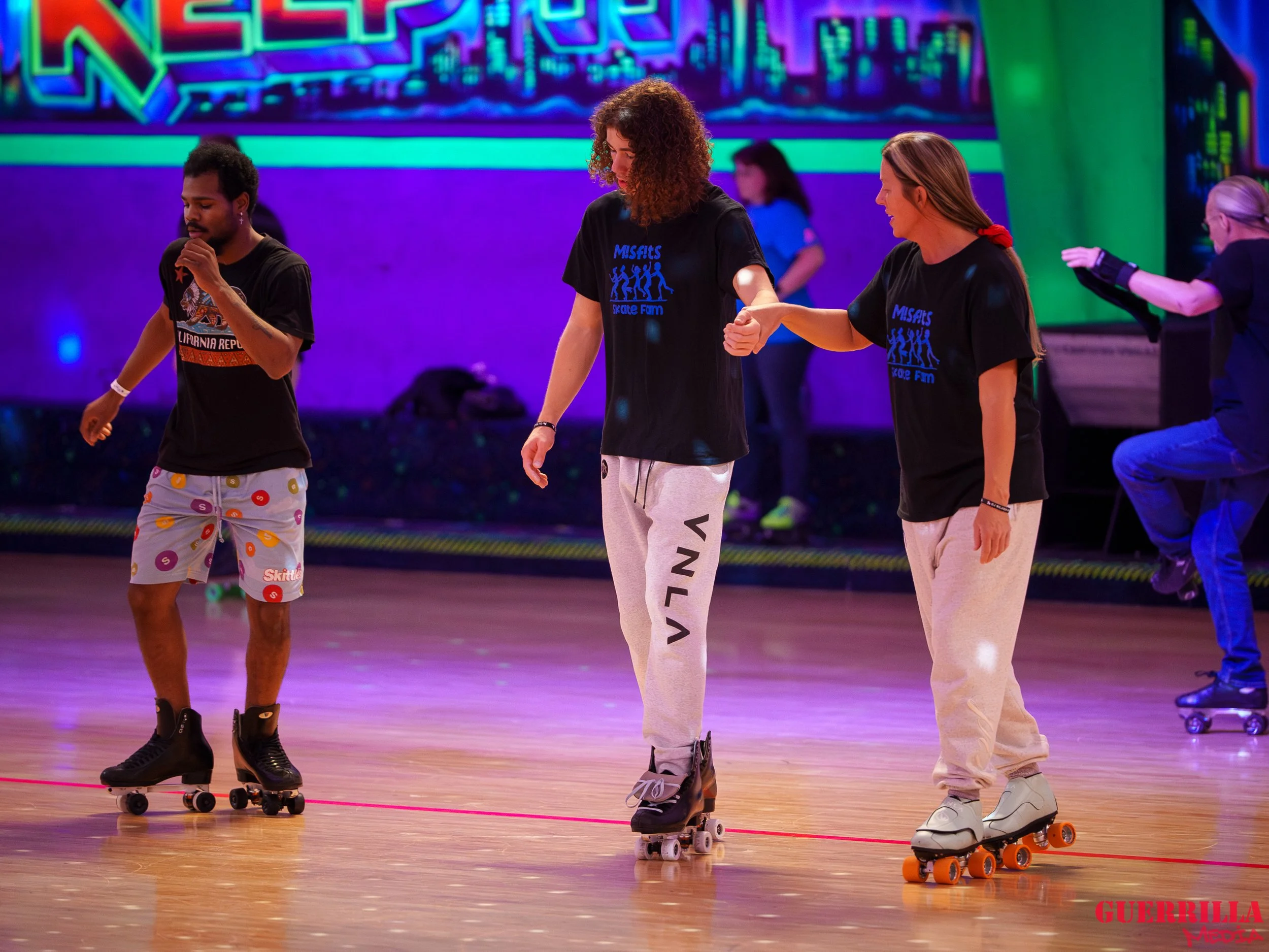 Three people roller skating on an indoor rink, with two women holding hands and a man skating nearby. The background features colorful neon lights and a large digital display.