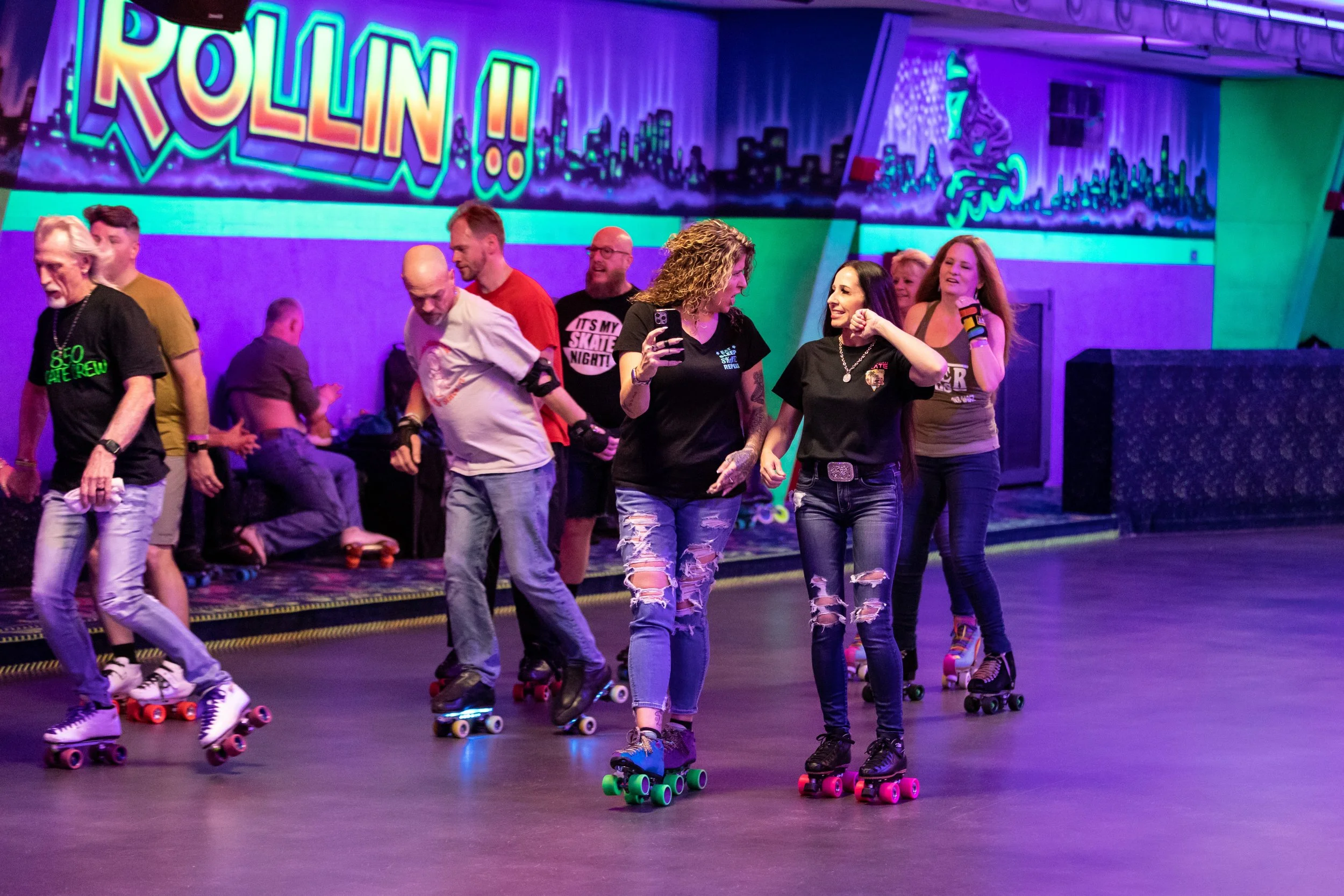 People roller skating in an indoor rink with neon purple lighting and a colorful cityscape mural in the background. The sign says "ROLLIN!!" with a city skyline and roller skate graphic.