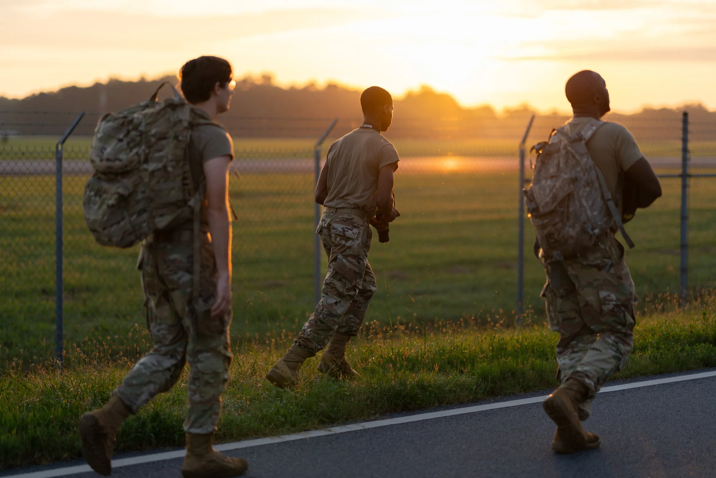 Three soldiers in camouflage uniforms walking along a road at sunset, carrying backpacks, near a fence.