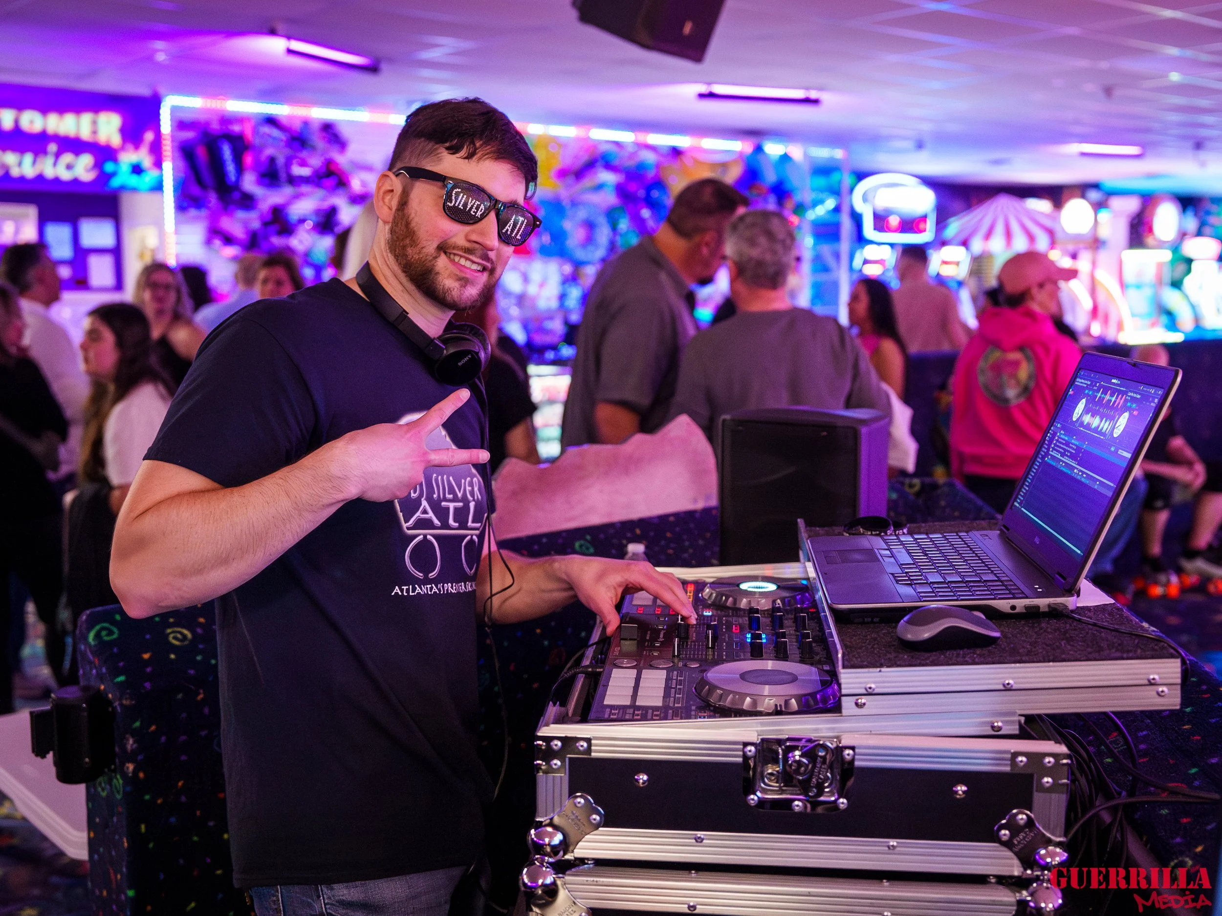 A DJ wearing sunglasses and a black T-shirt, smiling and flashing a peace sign, standing behind a DJ setup with a laptop and mixer in a neon-lit, crowded arcade or club.
