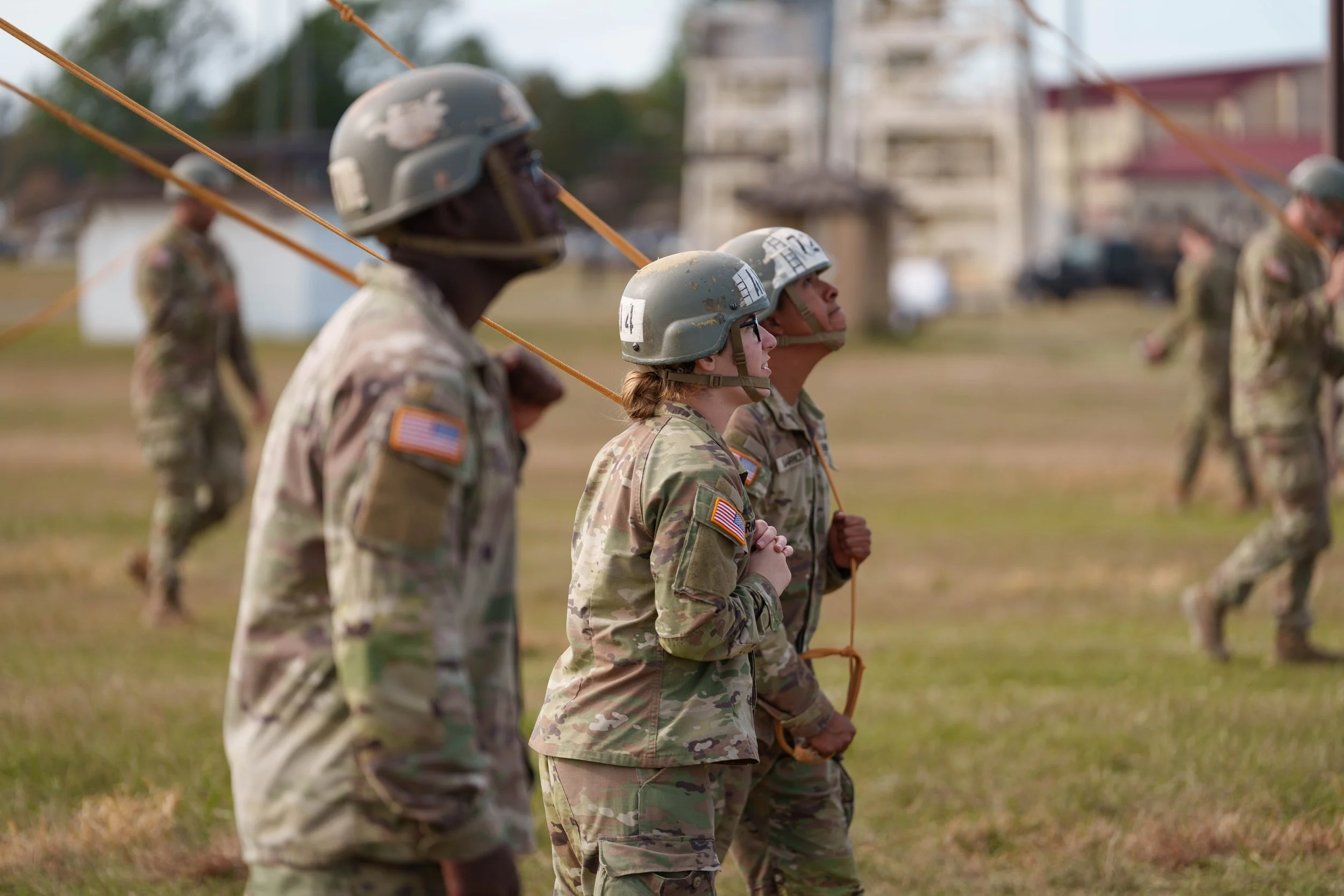 A group of soldiers in camouflage uniforms and helmets standing in a field during a military training exercise, holding on to ropes with serious expressions.