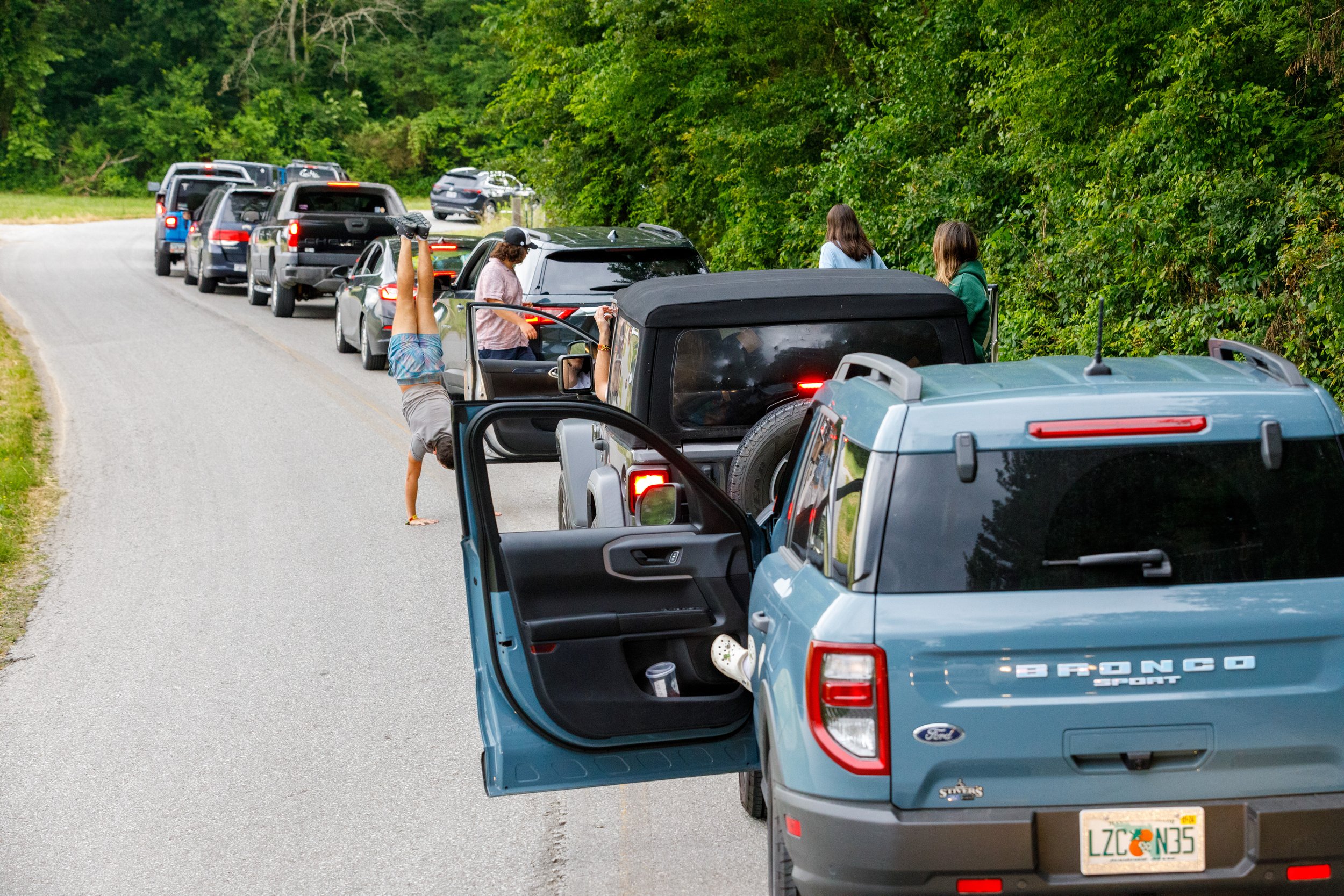 Line of parked cars on the side of a rural road with a group of people, some standing, chatting, and one person doing a handstand in front of the cars, surrounded by green trees.