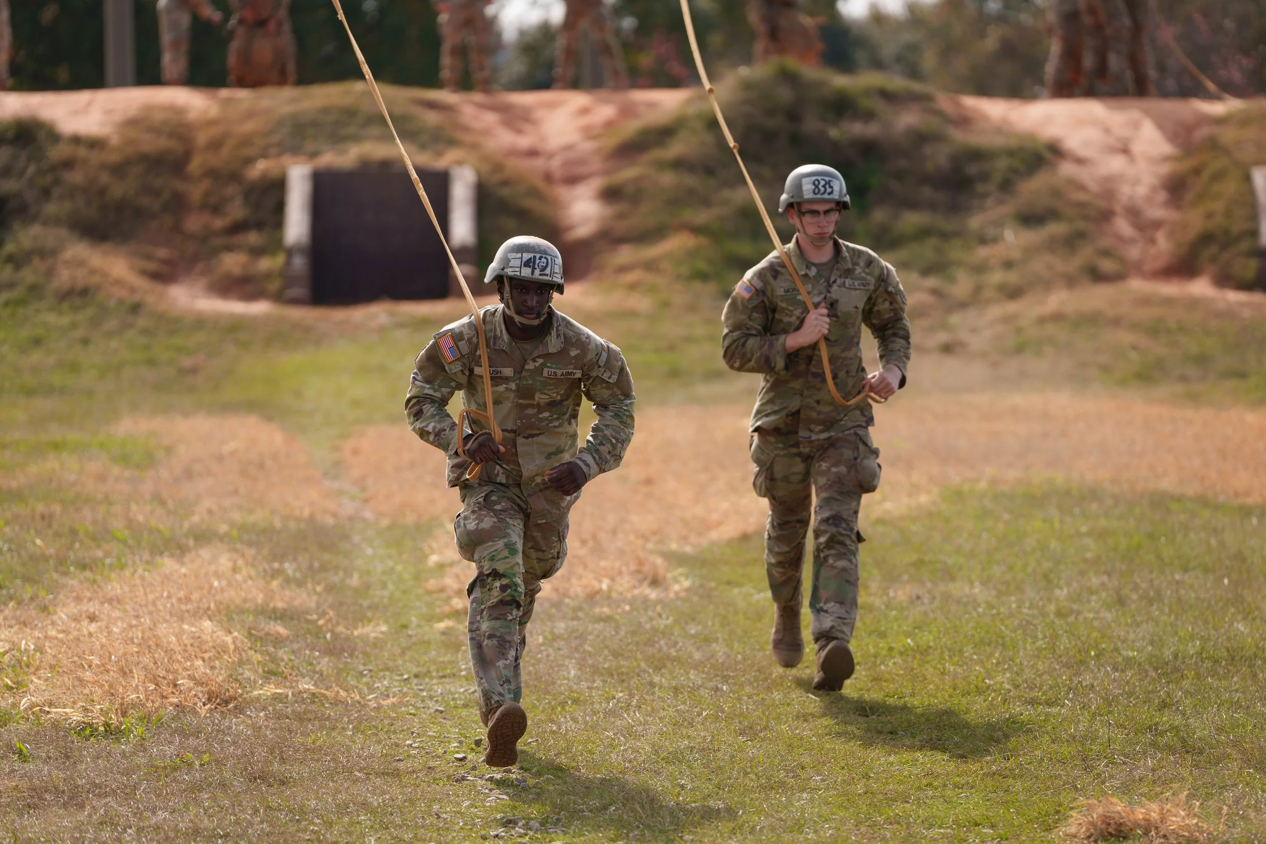 Two soldiers in camouflage uniforms running across a grassy field, holding ropes, with trees in the background.