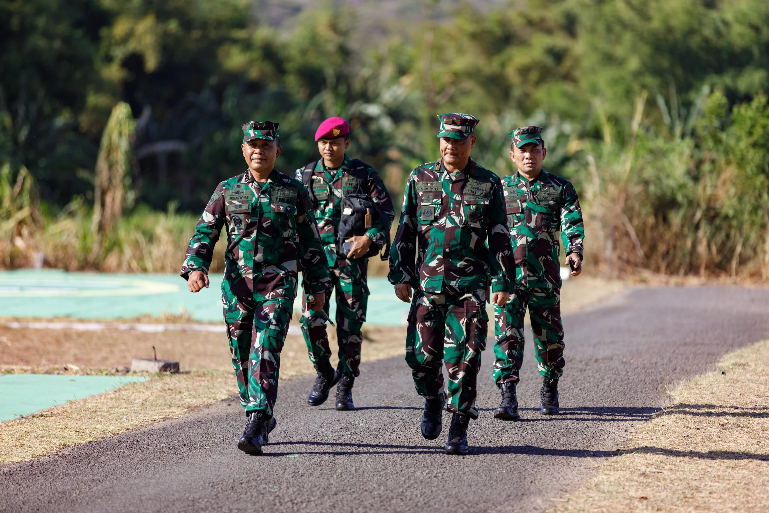 Five military personnel marching on a paved road surrounded by greenery.