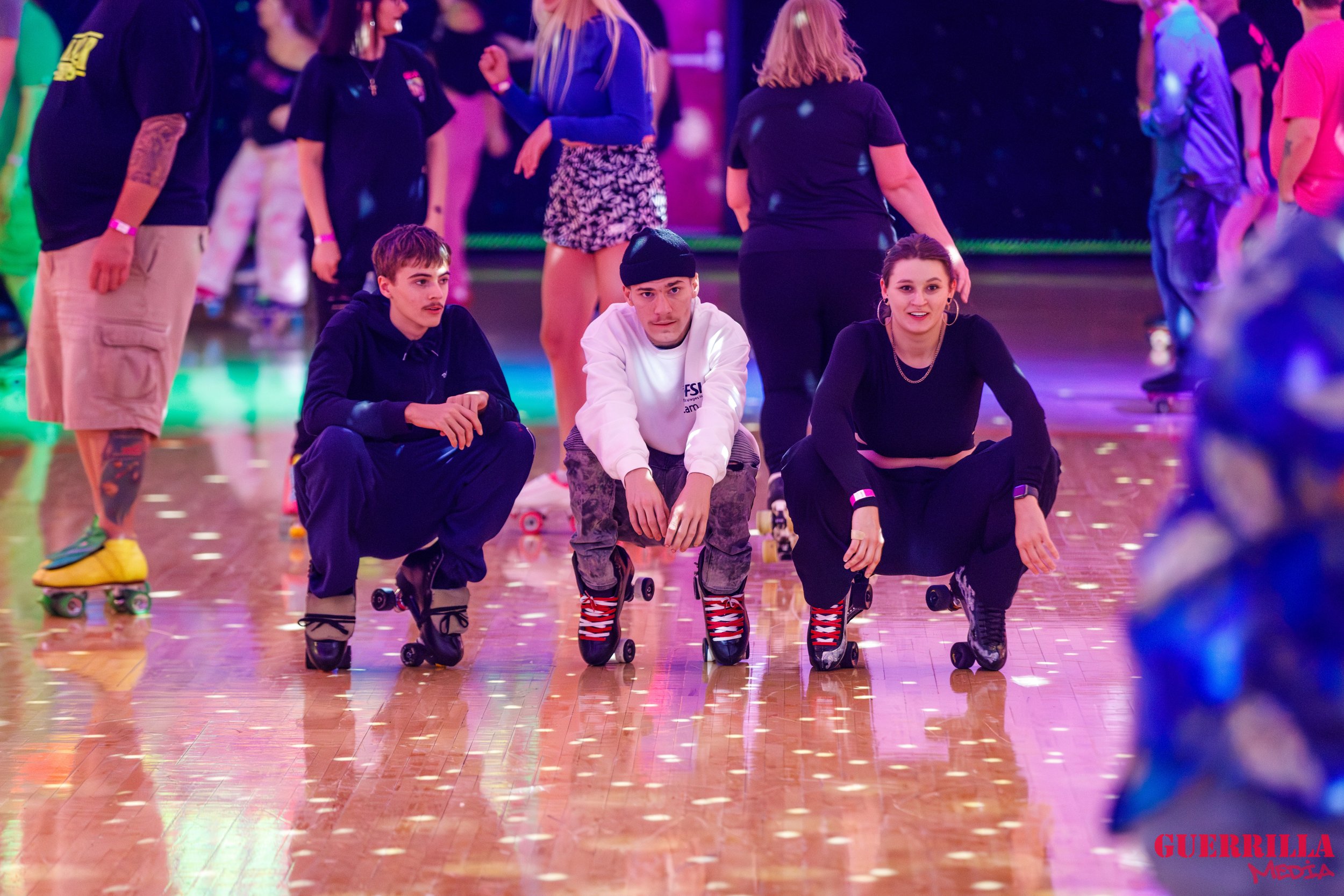 Three teenagers in roller skates crouching on a polished wooden floor at an indoor roller skating rink, with colorful lights and other skaters in the background.