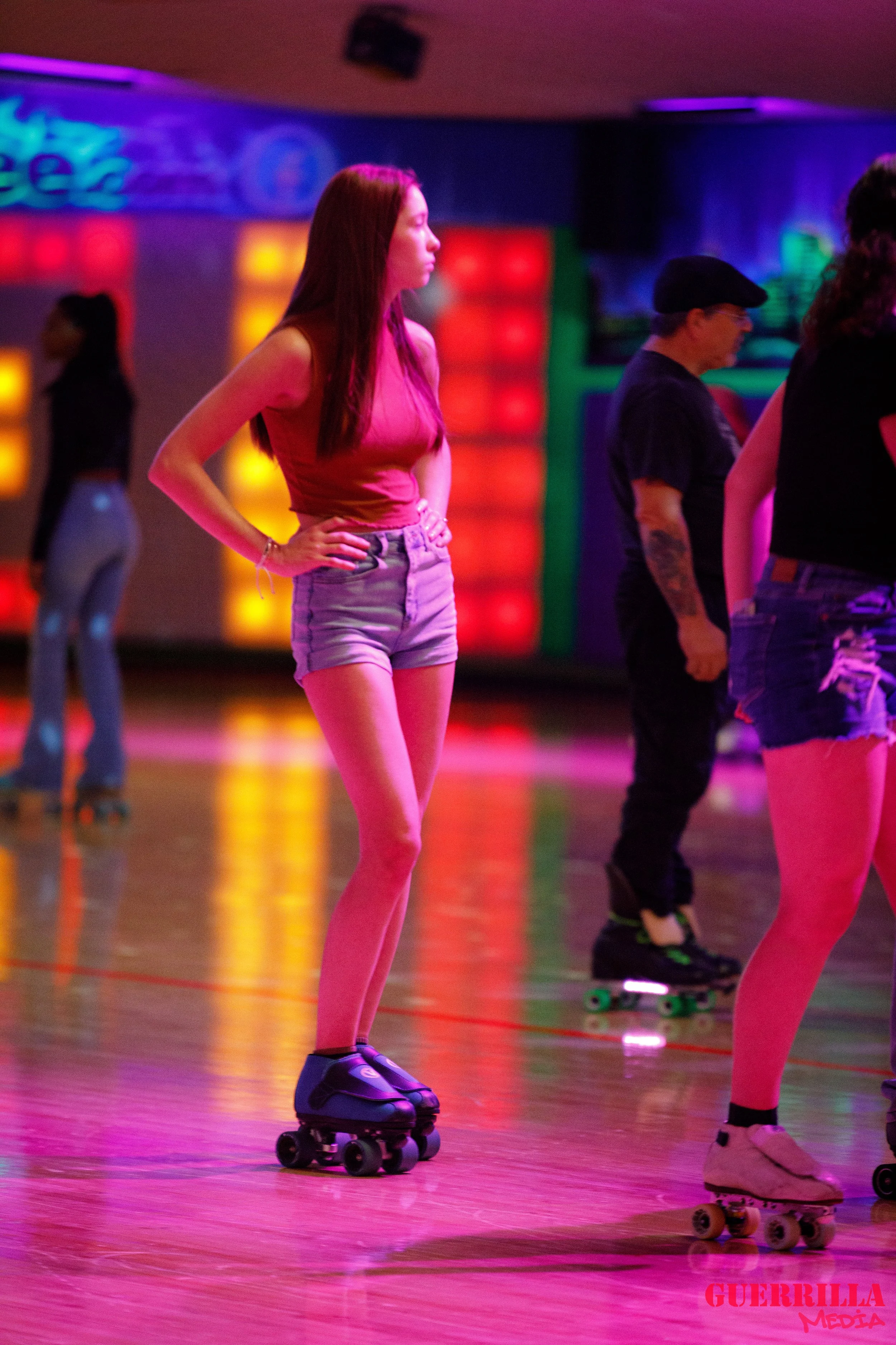 A young woman with long red hair roller skating in a nightclub with neon lights, wearing a pink sleeveless top and denim shorts.