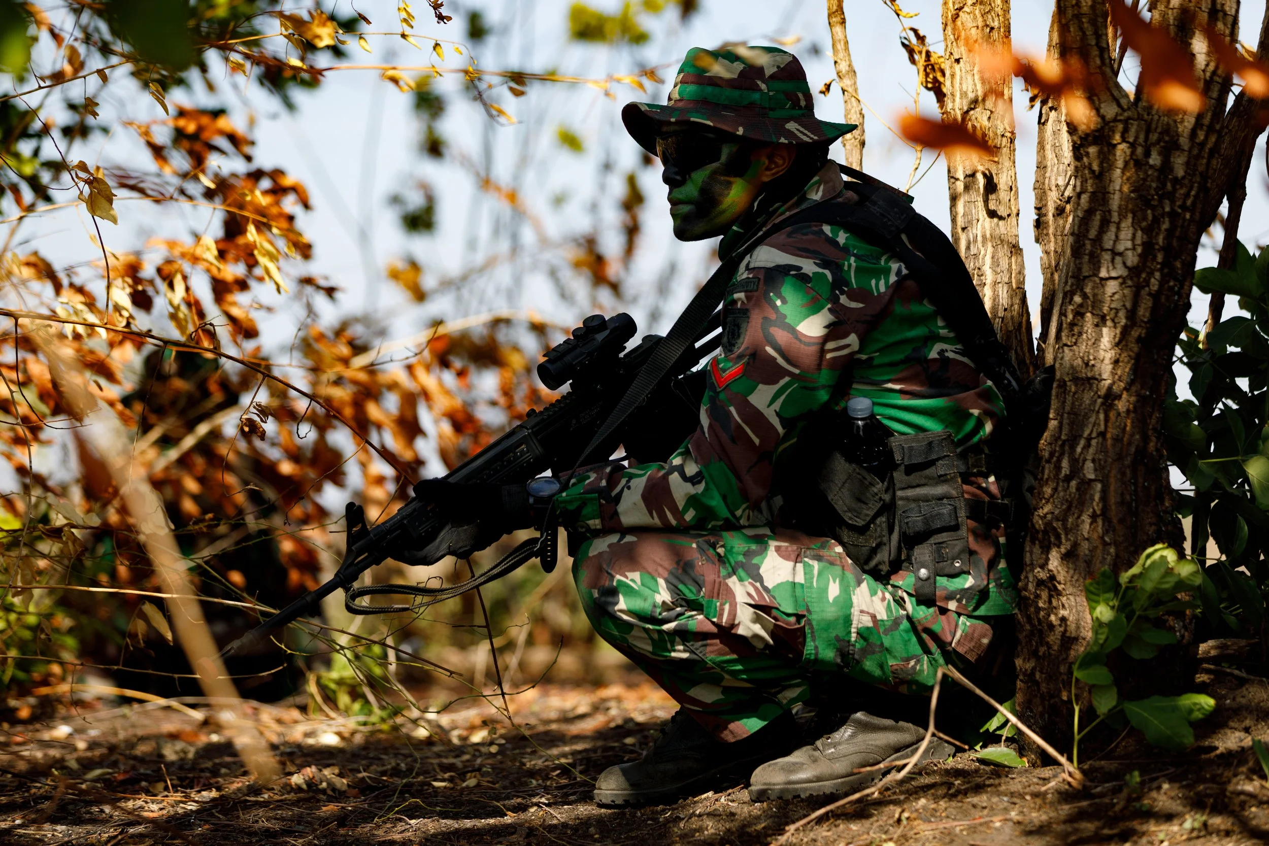 Soldier in camouflage uniform crouching behind a tree with a rifle, amid an outdoor woodland setting with brown leaves.