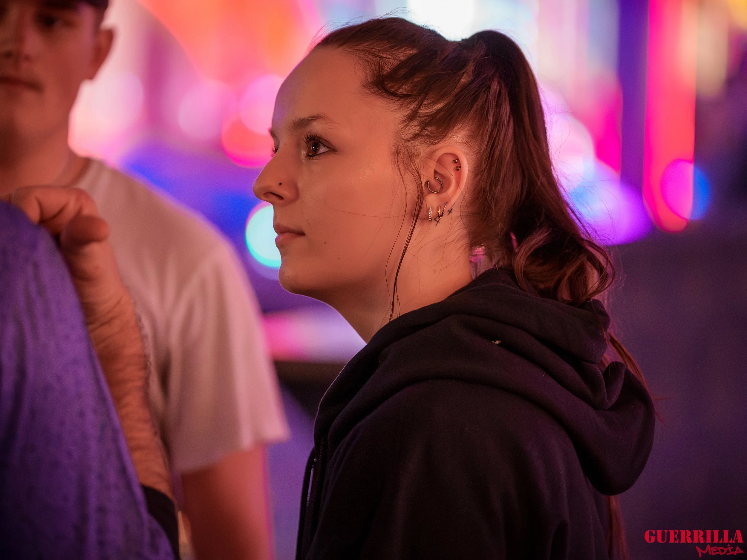 A young woman with multiple ear piercings, wearing a black hoodie, looking to the right in front of colorful and blurry neon lights.