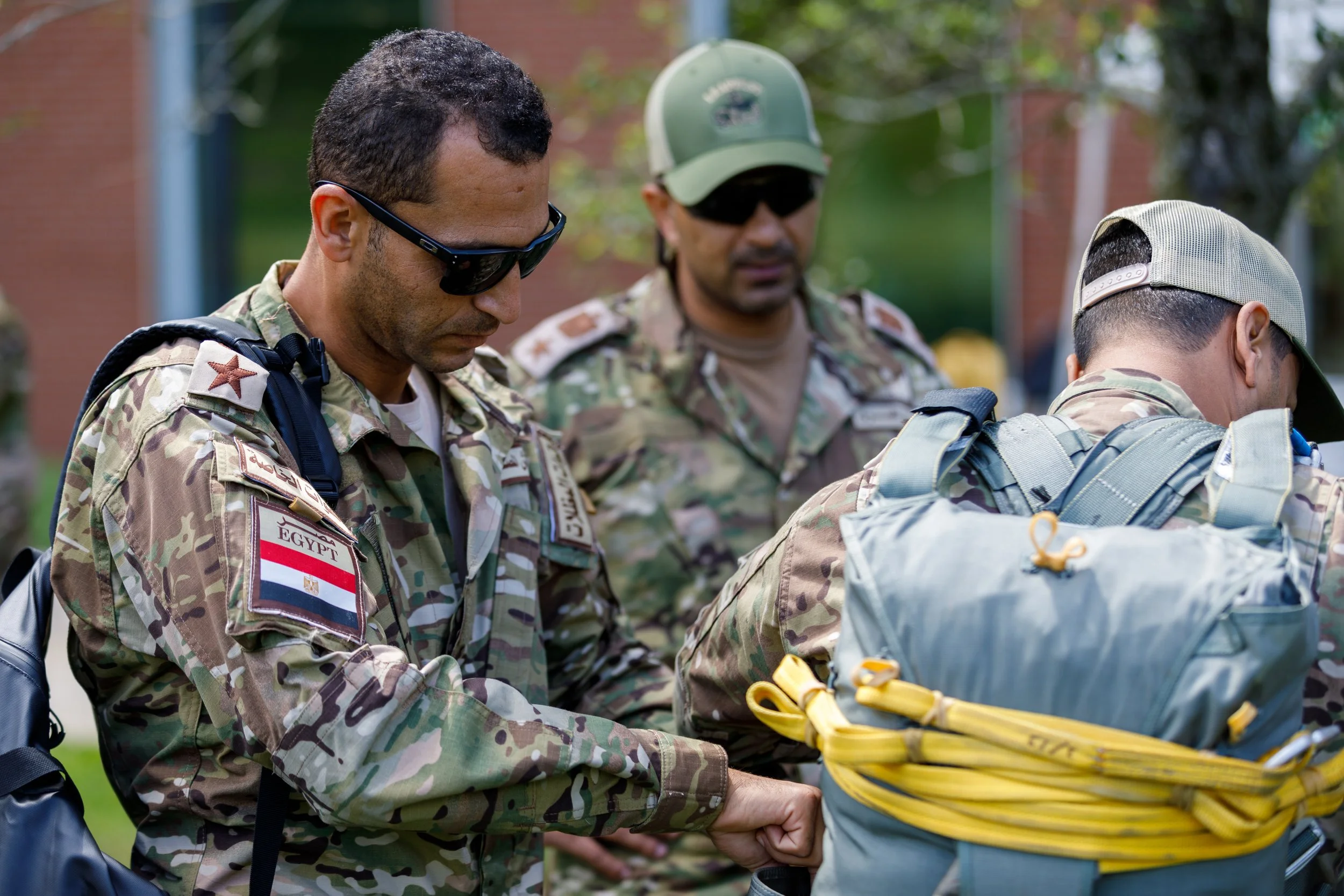 Three military personnel in camouflage uniforms, two wearing sunglasses and caps, one with a backpack, gathered outdoors in front of greenery and a brick building.