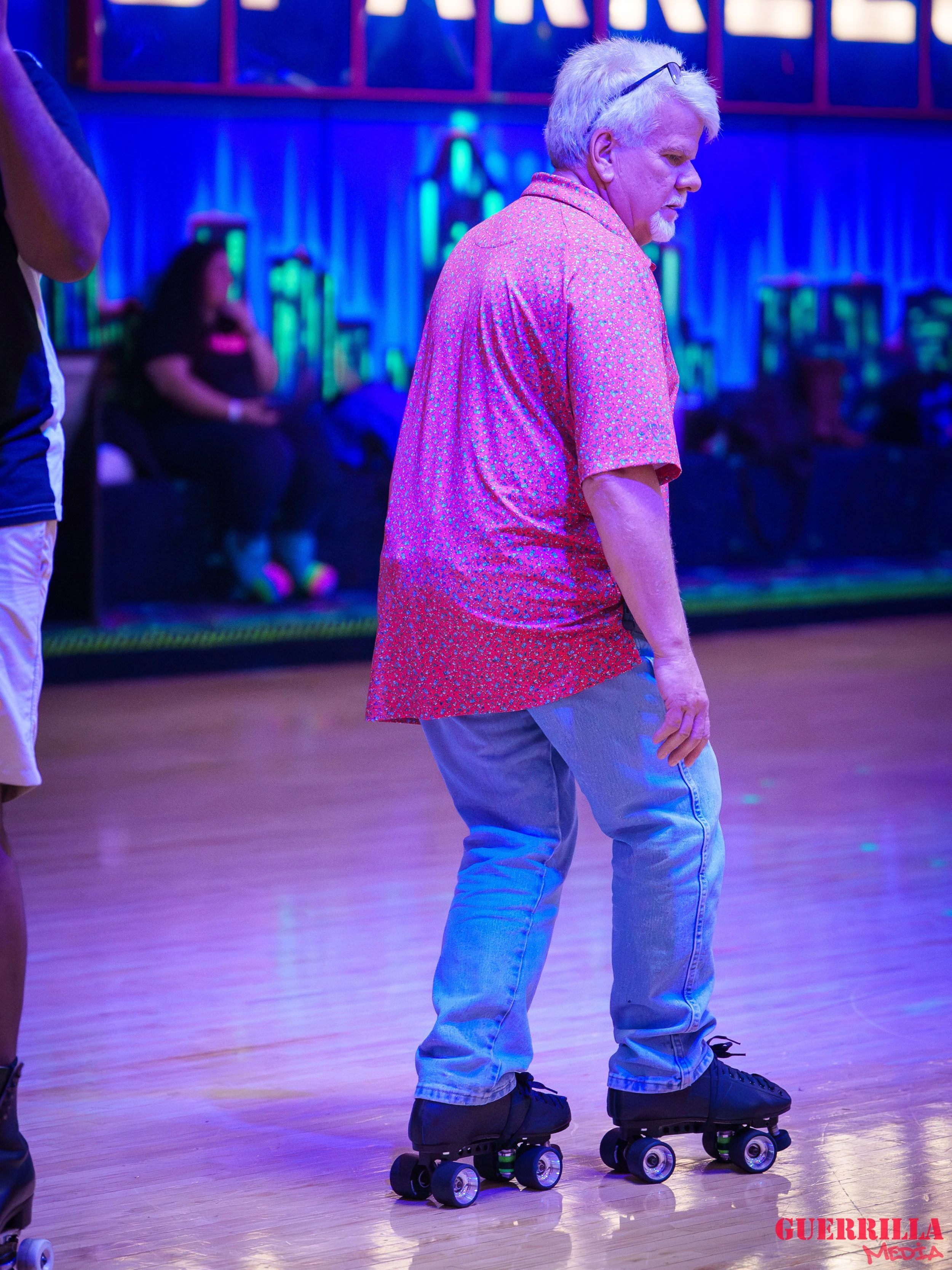 An elderly man with white hair and glasses is roller skating in a neon-lit indoor roller rink, wearing a red patterned shirt and light blue jeans.