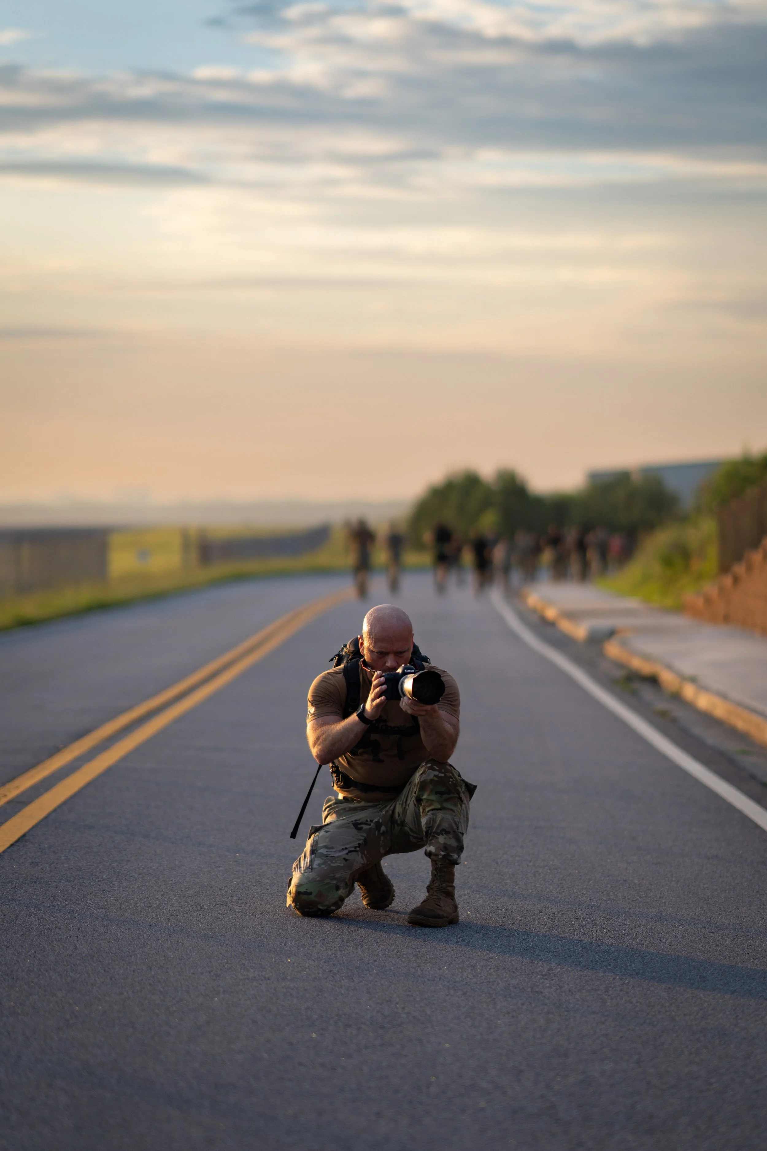 A person in military camouflage kneeling on the road, photographing a group of people in the distance at sunset.
