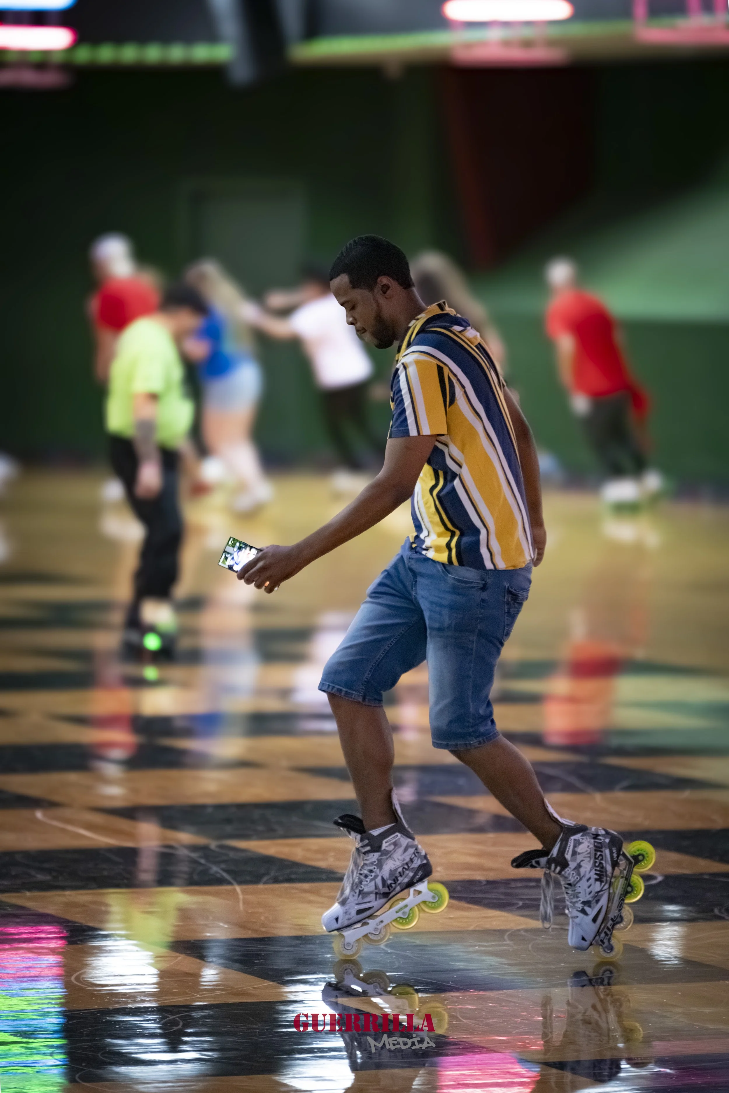A man rollerblading on a checkered floor while looking at his phone, with other people in the background at an indoor roller rink.