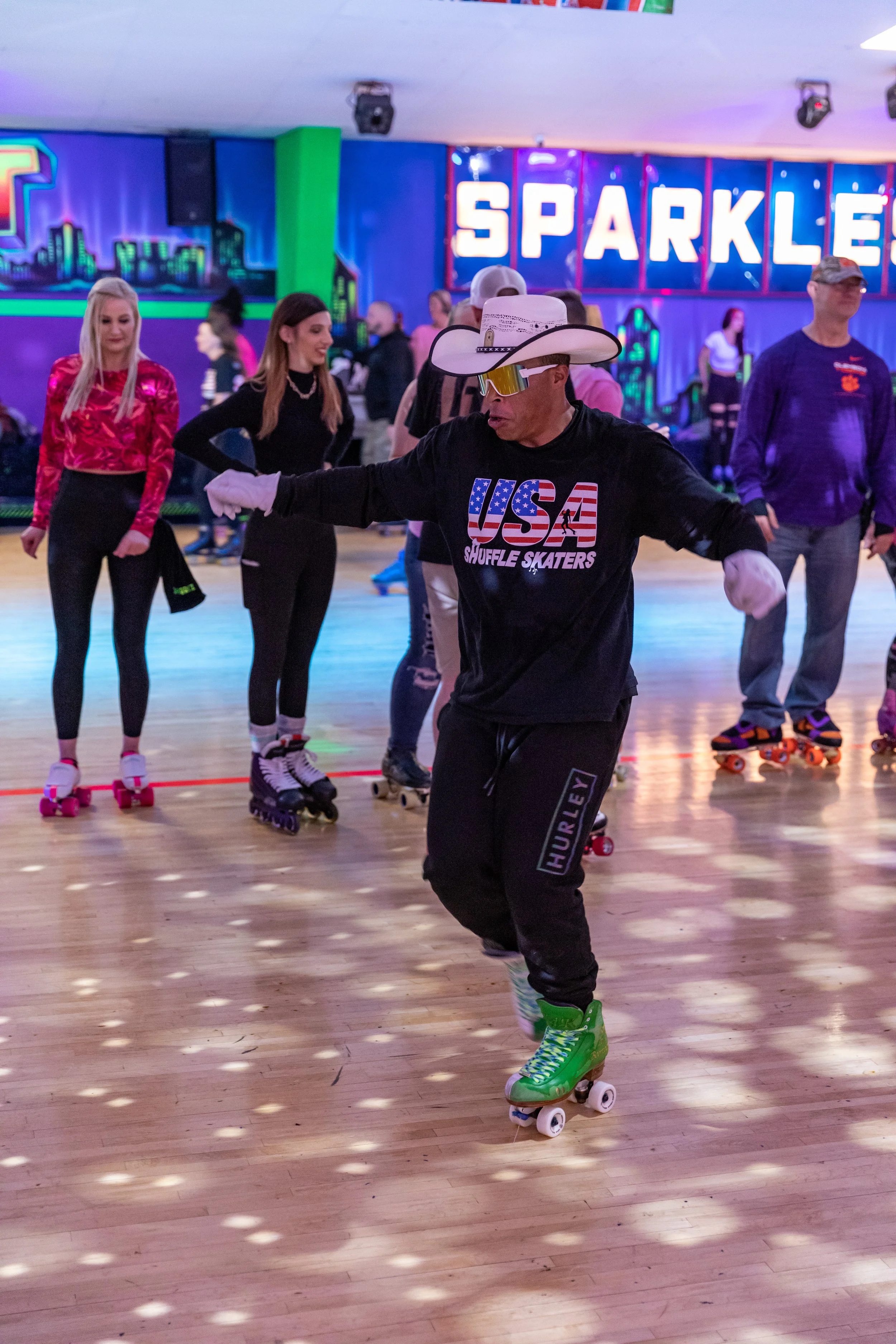 A man roller skating indoors on a wooden floor, wearing a cowboy hat, sunglasses, a black USA Shuffle Skaters sweatshirt, and green roller skates, with several people in the background also skating and a large illuminated sign that says 'SPARKLE'.