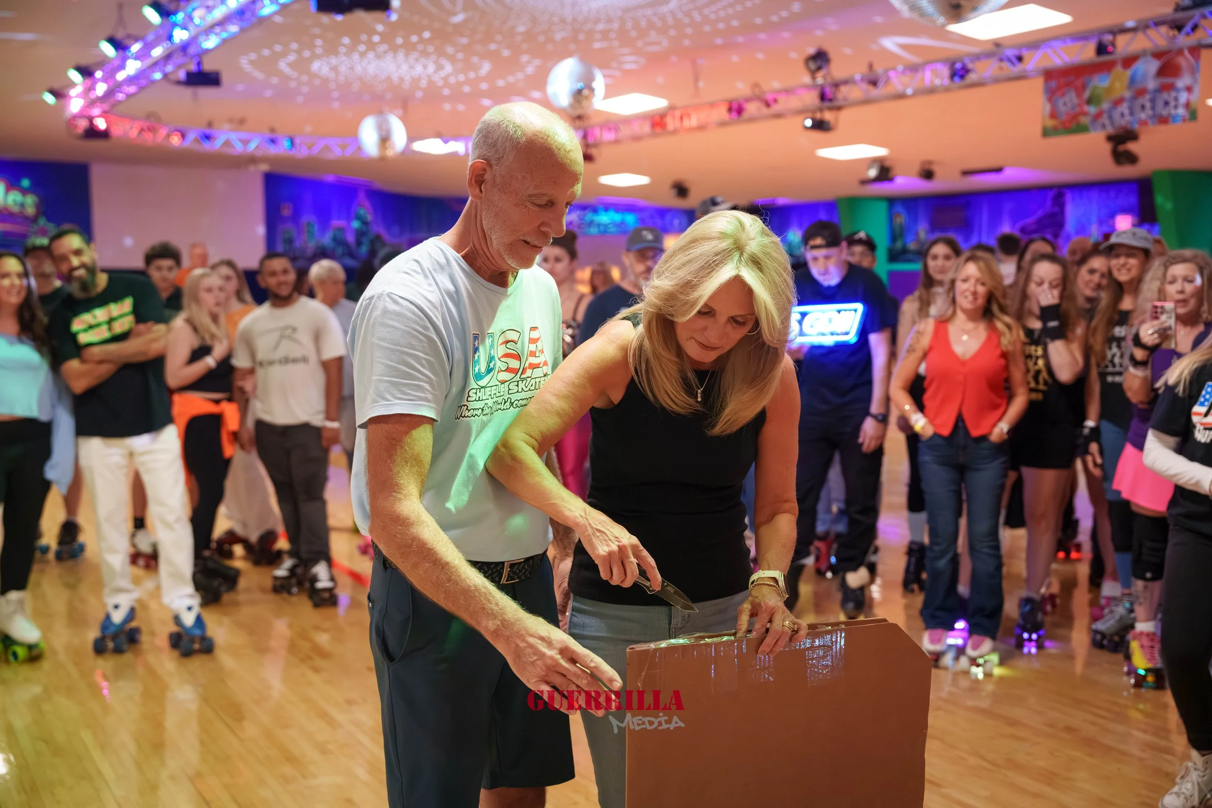 An elderly man and woman roller skate at an indoor roller disco, with a crowd watching, colorful lighting, and disco balls overhead.
