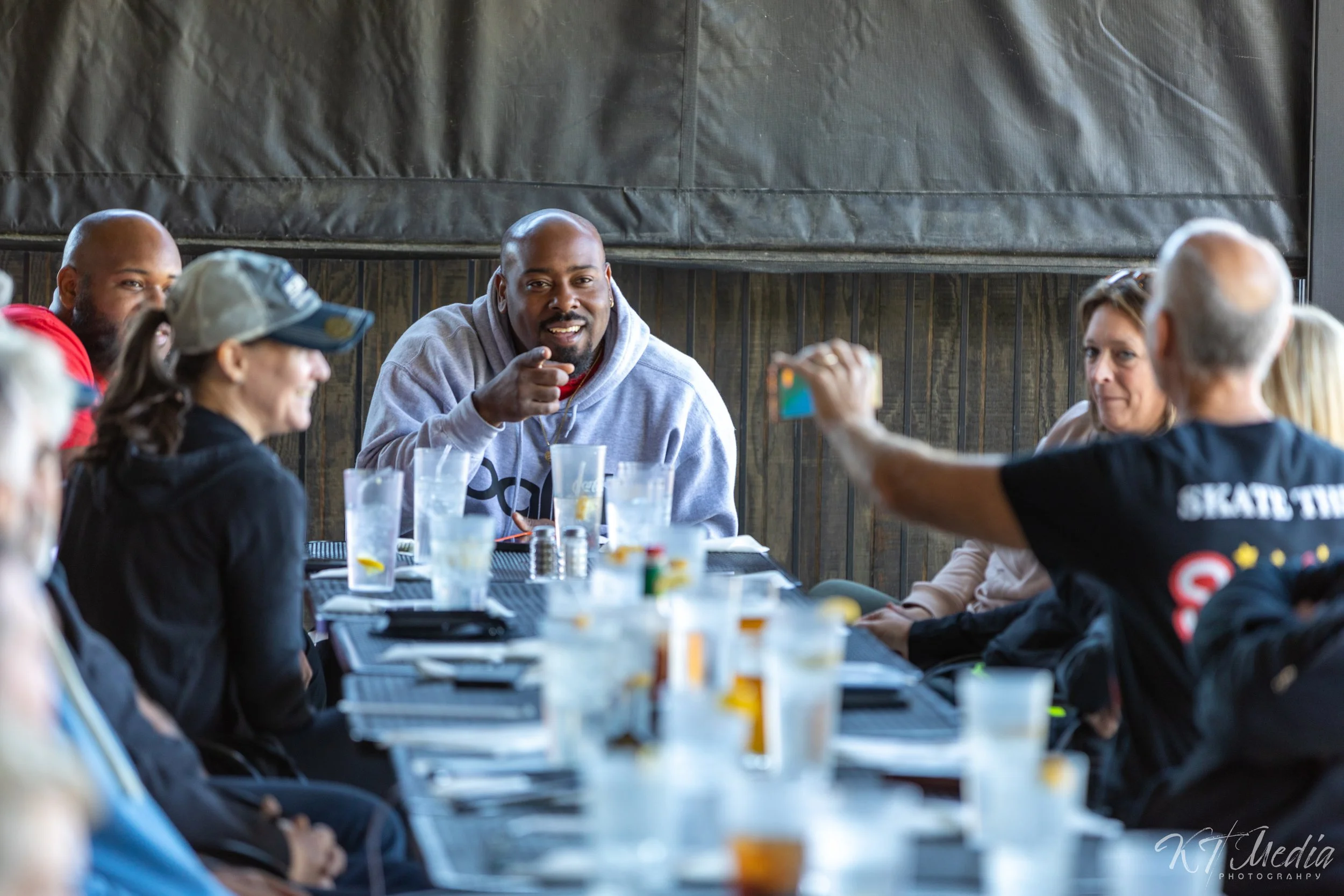 A group of people sitting around a table in a restaurant or cafe, engaged in a conversation. The table has glasses of drinks and snacks, with a man in a gray hoodie at the center smiling and pointing. Others are listening, some taking photos or video