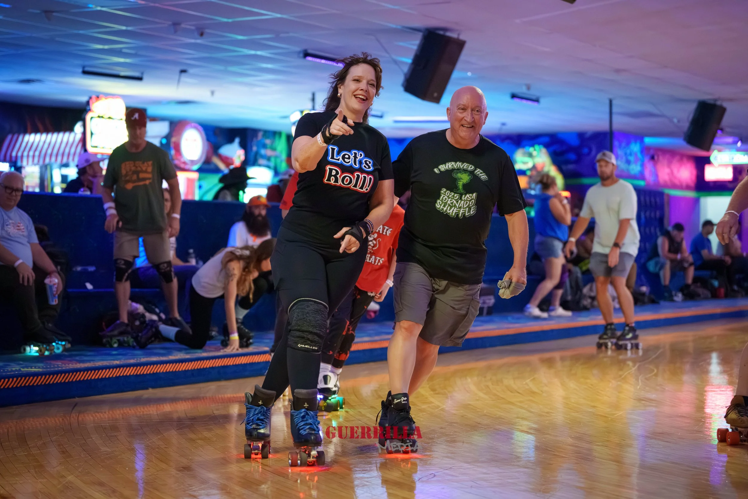 People roller skating at an indoor roller rink with colorful lights and neon signs in the background. Two women in the foreground are smiling and interacting, one wearing a black shirt with 'Let's Roll!' and the other wearing a black shirt with a gre