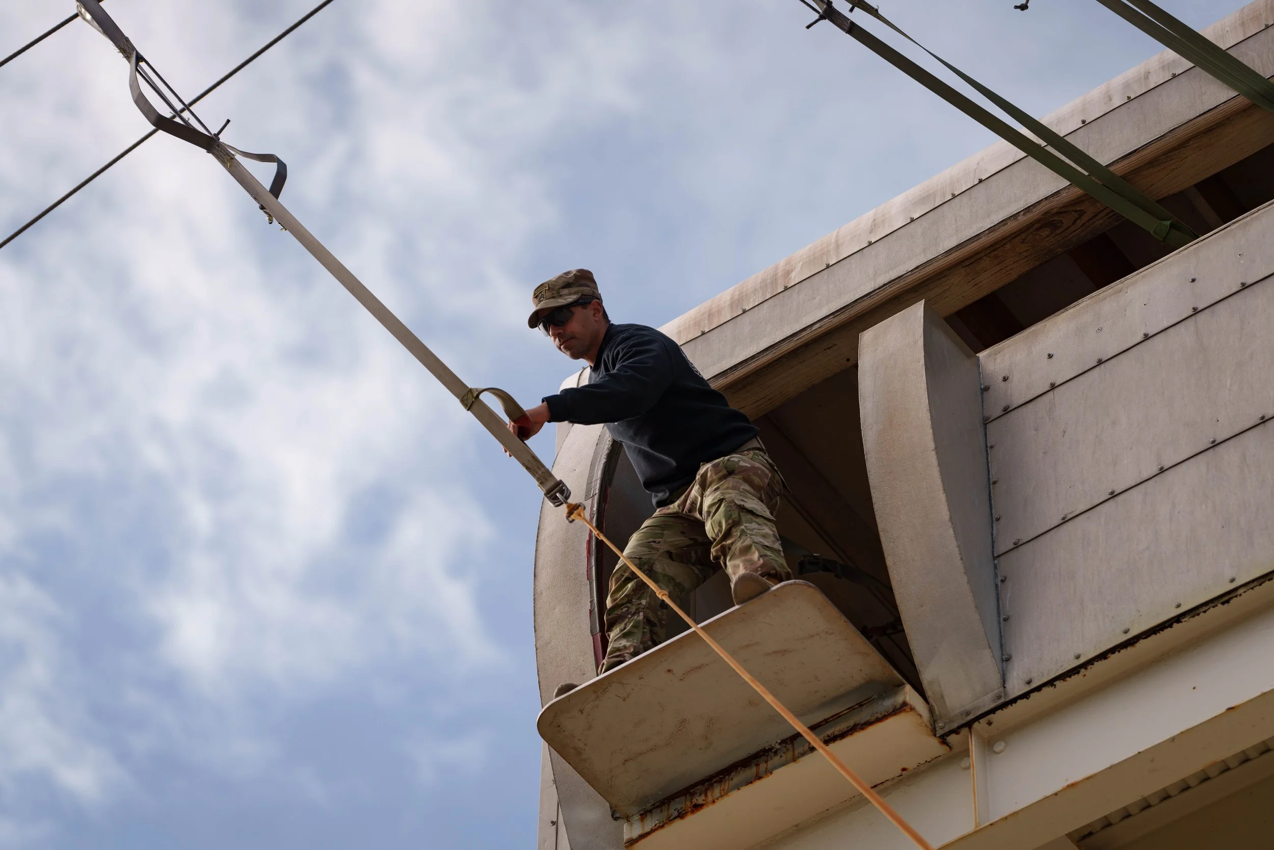 A man in military camouflage pants and a black jacket works on a construction site, standing on a small platform attached to the side of a building, holding a securing strap with a background of a cloudy sky.