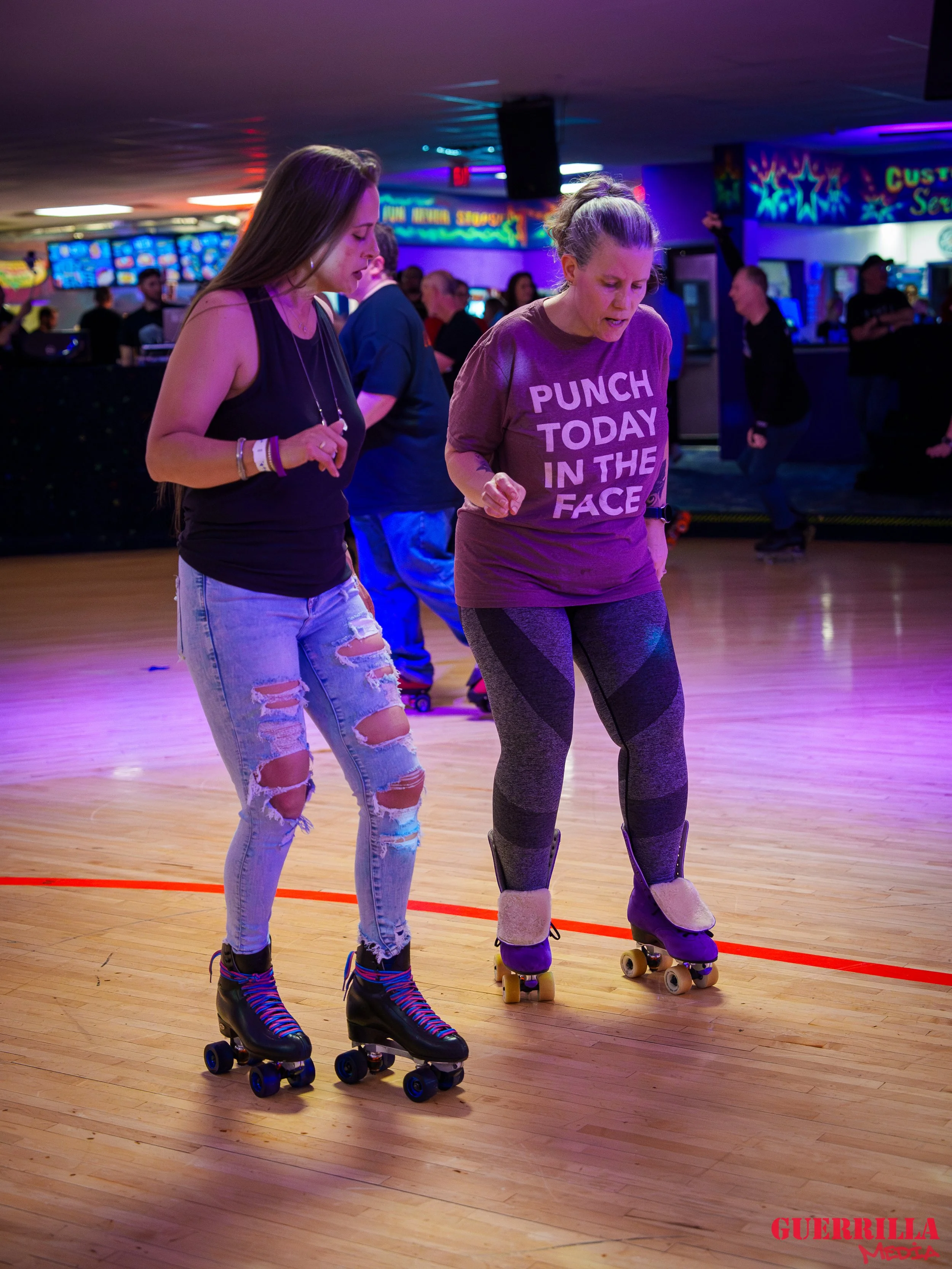 Two women roller skating in an indoor skating rink with colorful lights and other skaters in the background.