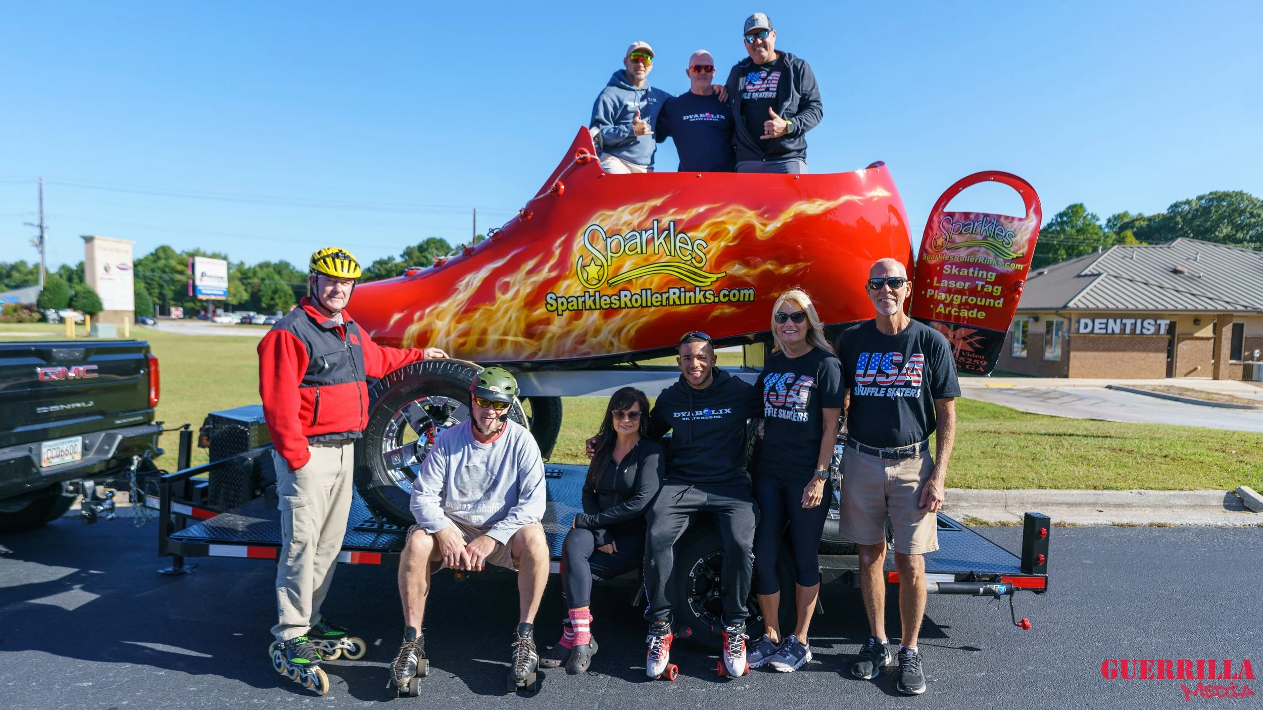 A group of nine people, some wearing roller skates, poses in front of a large, decorative roller coaster car labeled "Sparkles" with flames painted on it. The group is outdoors on a sunny day, and some are standing on skateboards or rollerblades.