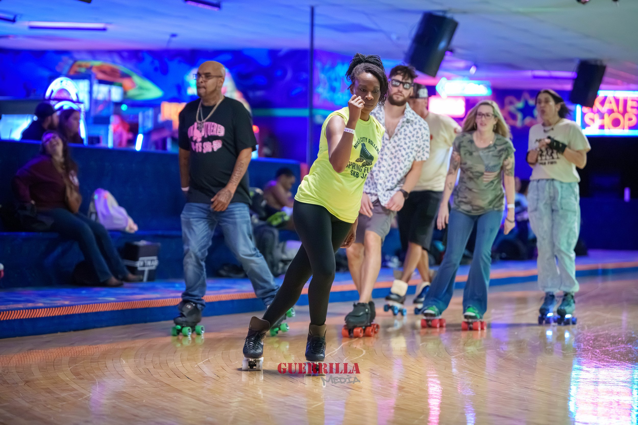 A group of people roller skating indoors under colorful neon lights, with some sitting on benches in the background.