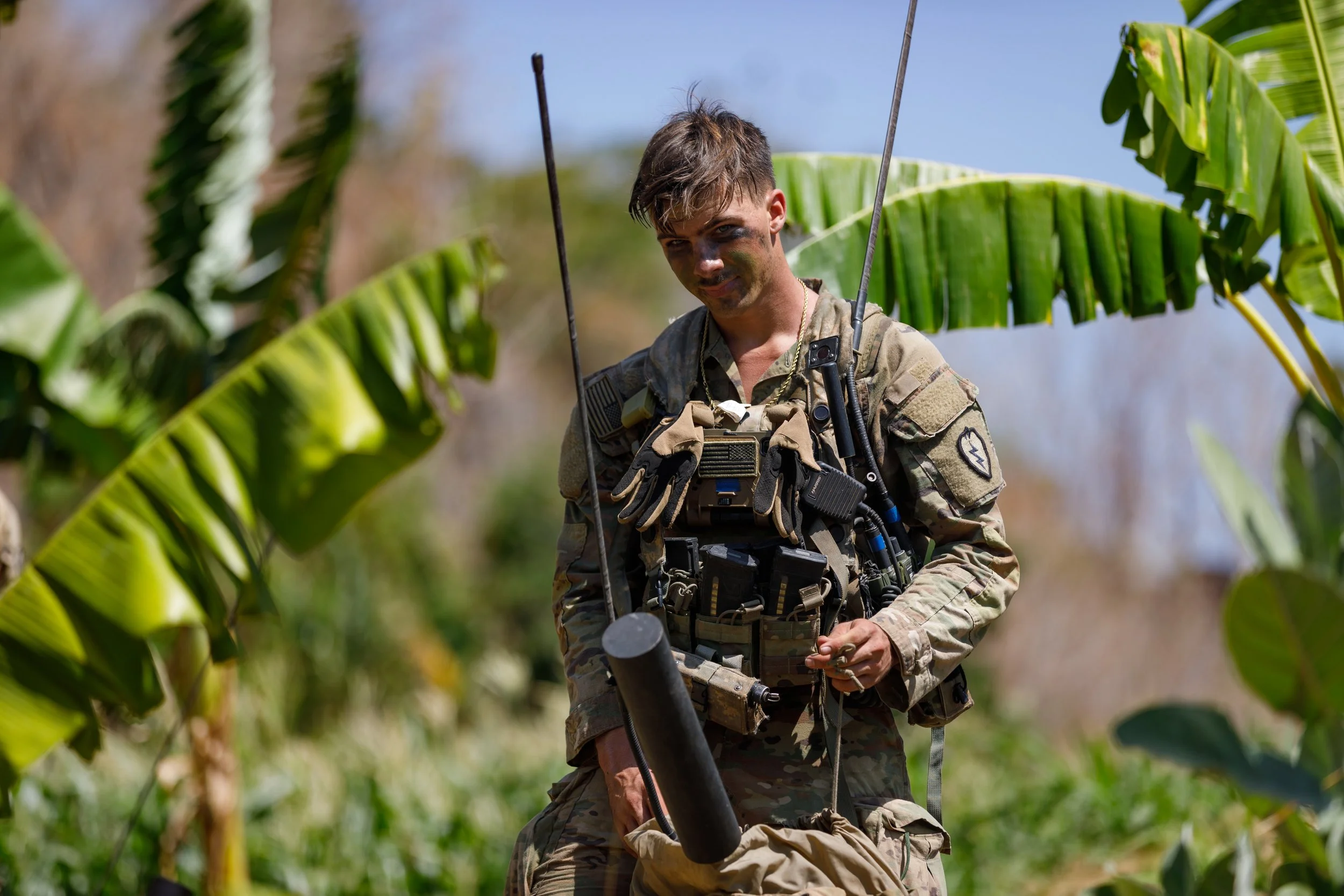 Soldier in camouflage uniform holding a device in a jungle with green banana leaves around.