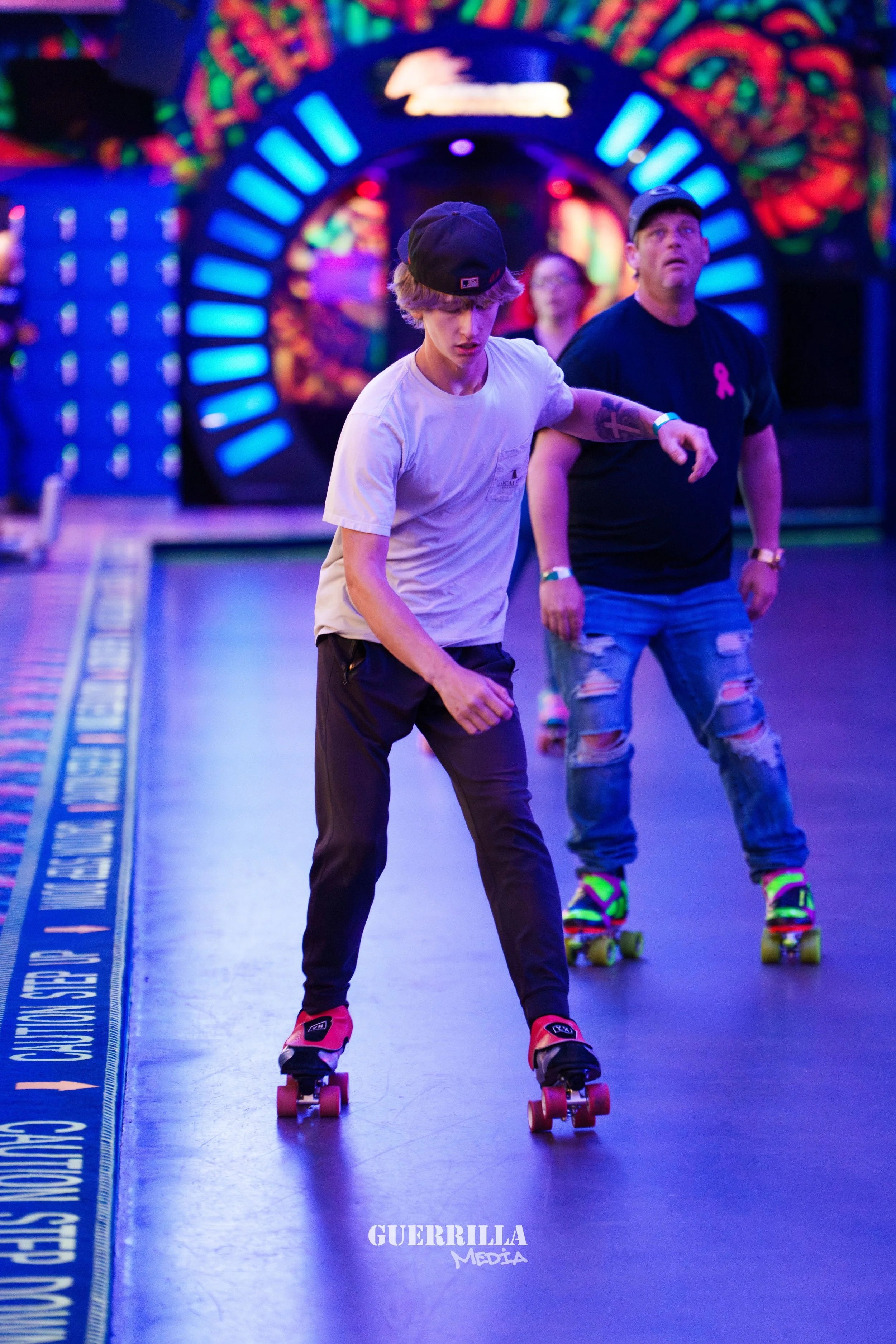 Teen boy roller skating at an indoor rink with a man in black t-shirt and ripped jeans, colorful neon lights in the background.