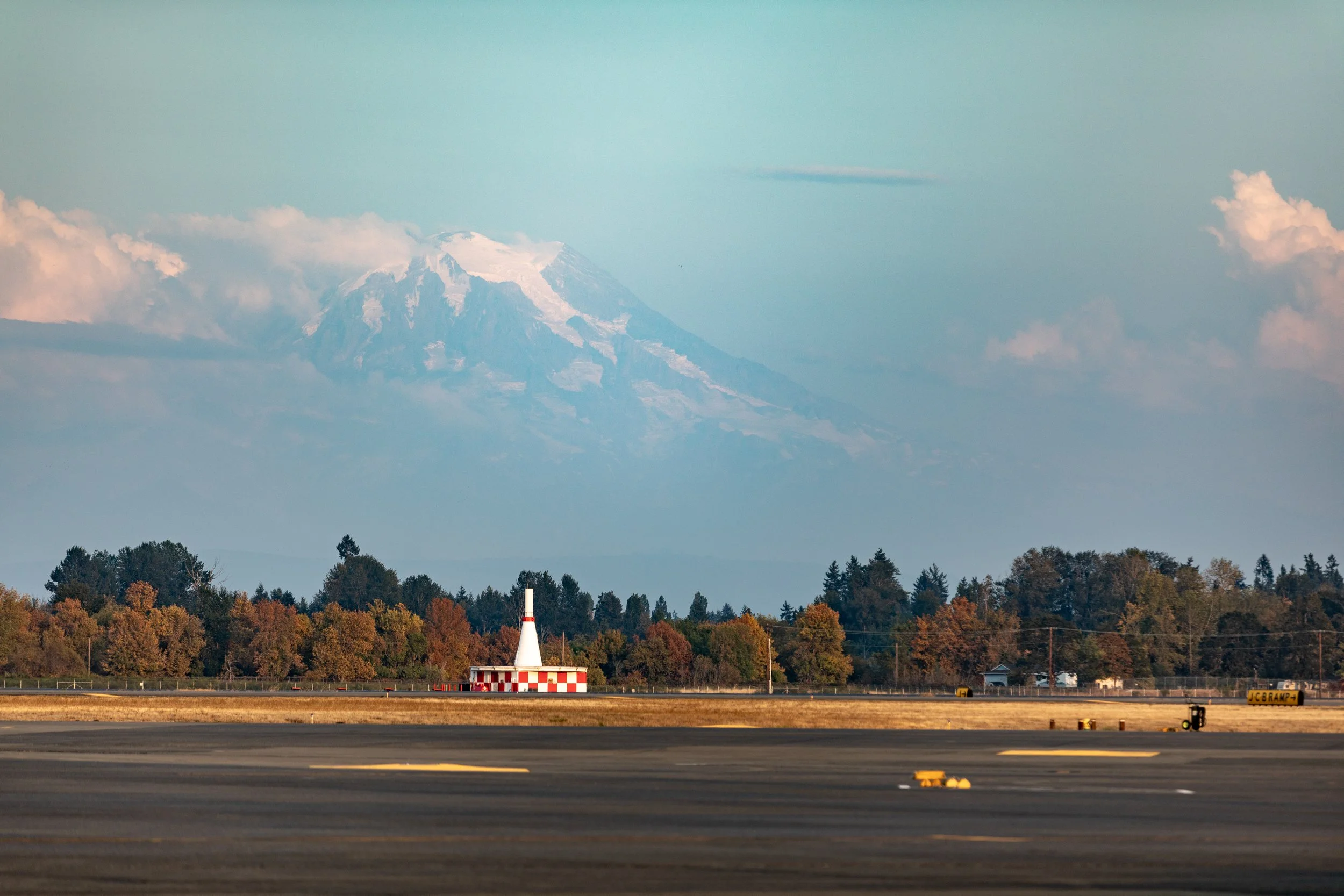 A mountain with snow on top in the distance, a row of trees with fall foliage, and an airport runway with a small building and a red checkerboard pattern, possibly a navigation aid or beacon.