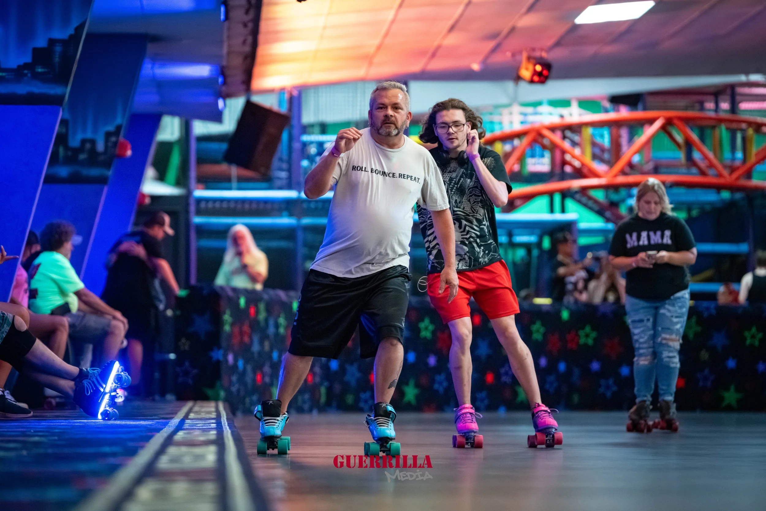 Two men roller skating inside an indoor roller rink with colorful lighting and a few people sitting and standing around.