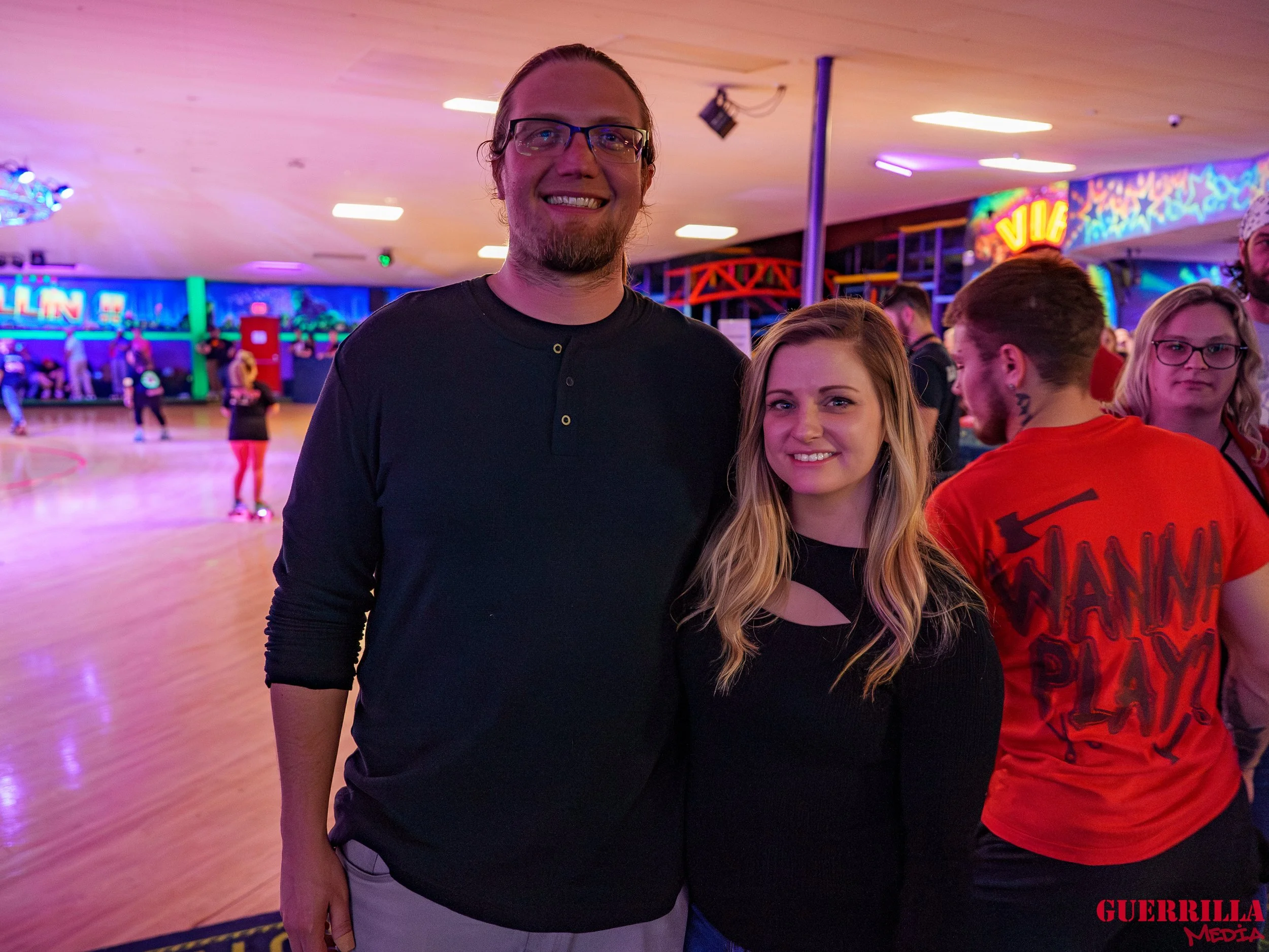 Two people, a man and a woman, standing together in a roller skating rink with colorful neon lights and other skaters in the background.