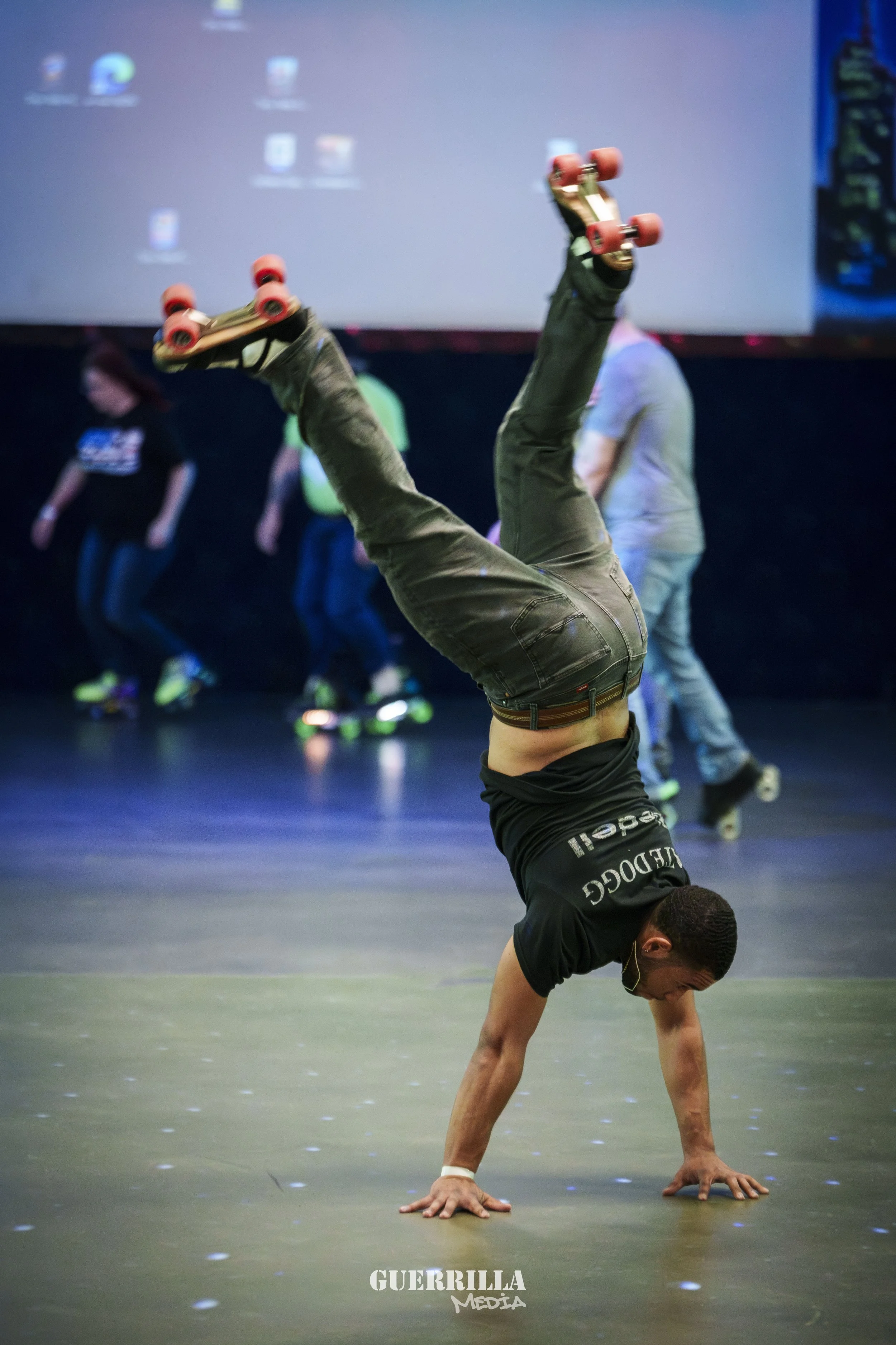 A man performing a handstand on roller skates in a dark room, with people in the background, and a large screen on the wall.