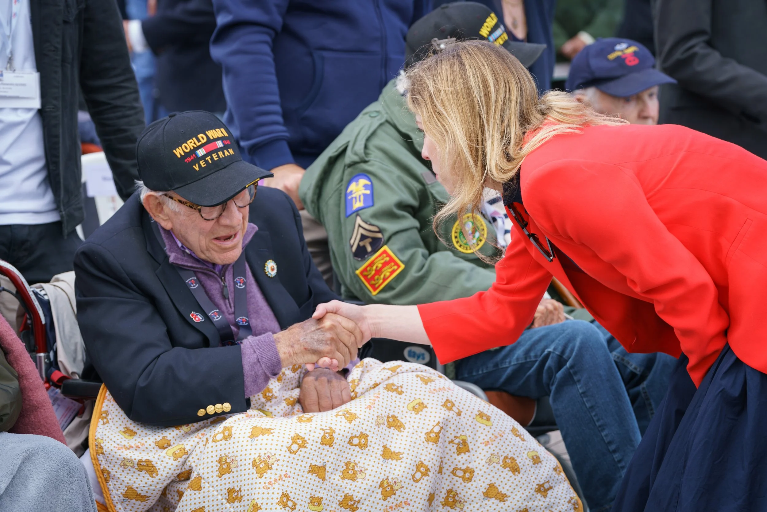 A young woman in a red blazer shakes hands with an elderly man in a wheelchair, who is wearing a Veterans cap and a dark blazer, surrounded by other people, some wearing military patches.